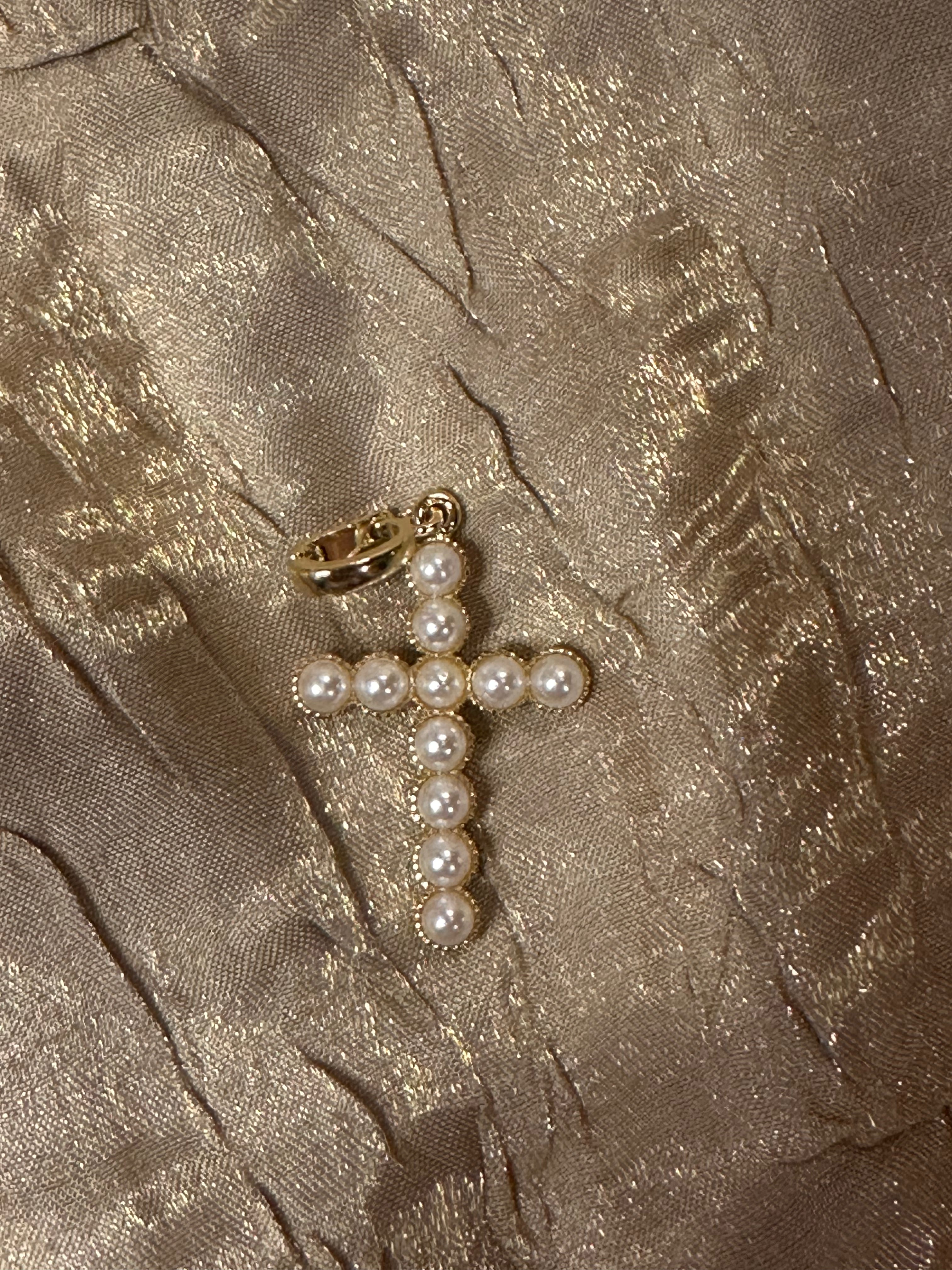 A gold cross pendant embellished with white faux pearls, resting on a textured, shimmering champagne-colored fabric background.