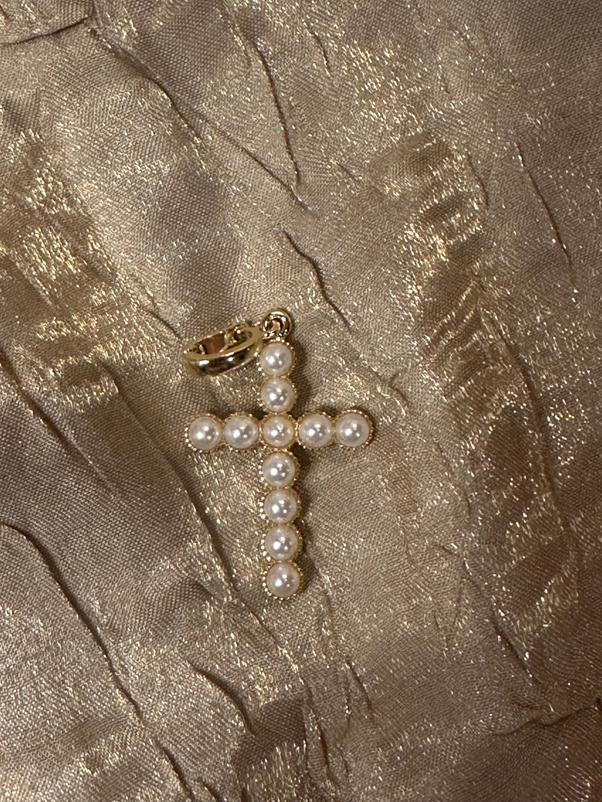 A gold cross pendant embellished with white faux pearls, resting on a textured, shimmering champagne-colored fabric background.