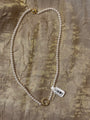 A white pearl necklace with gold-toned metal clasp details, laid flat on a textured gold fabric background with a white barcode tag attached.