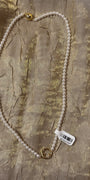 A white pearl necklace with gold-toned metal clasp details, laid flat on a textured gold fabric background with a white barcode tag attached.