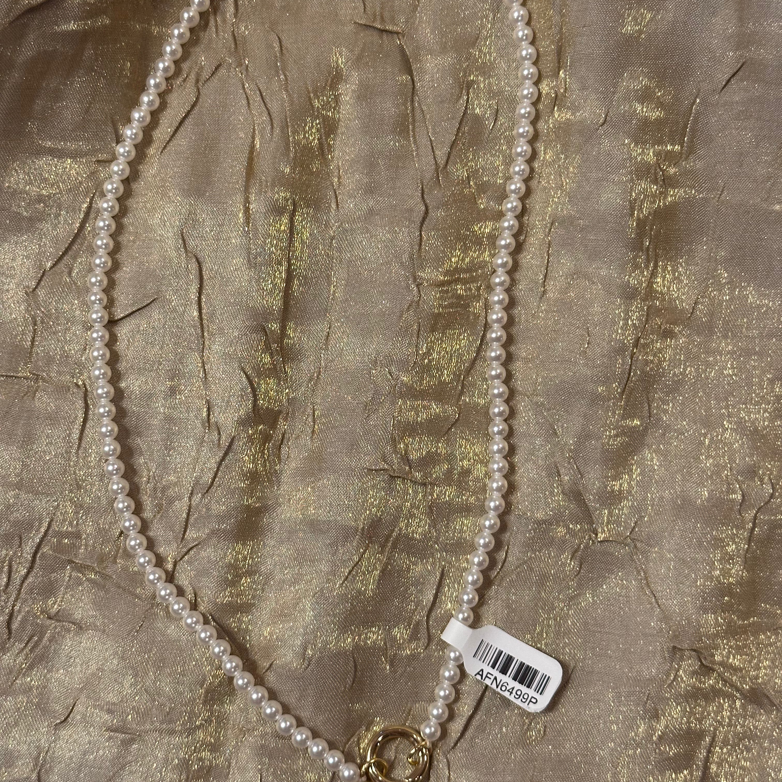 A white pearl necklace with gold-toned metal clasp details, laid flat on a textured gold fabric background with a white barcode tag attached.