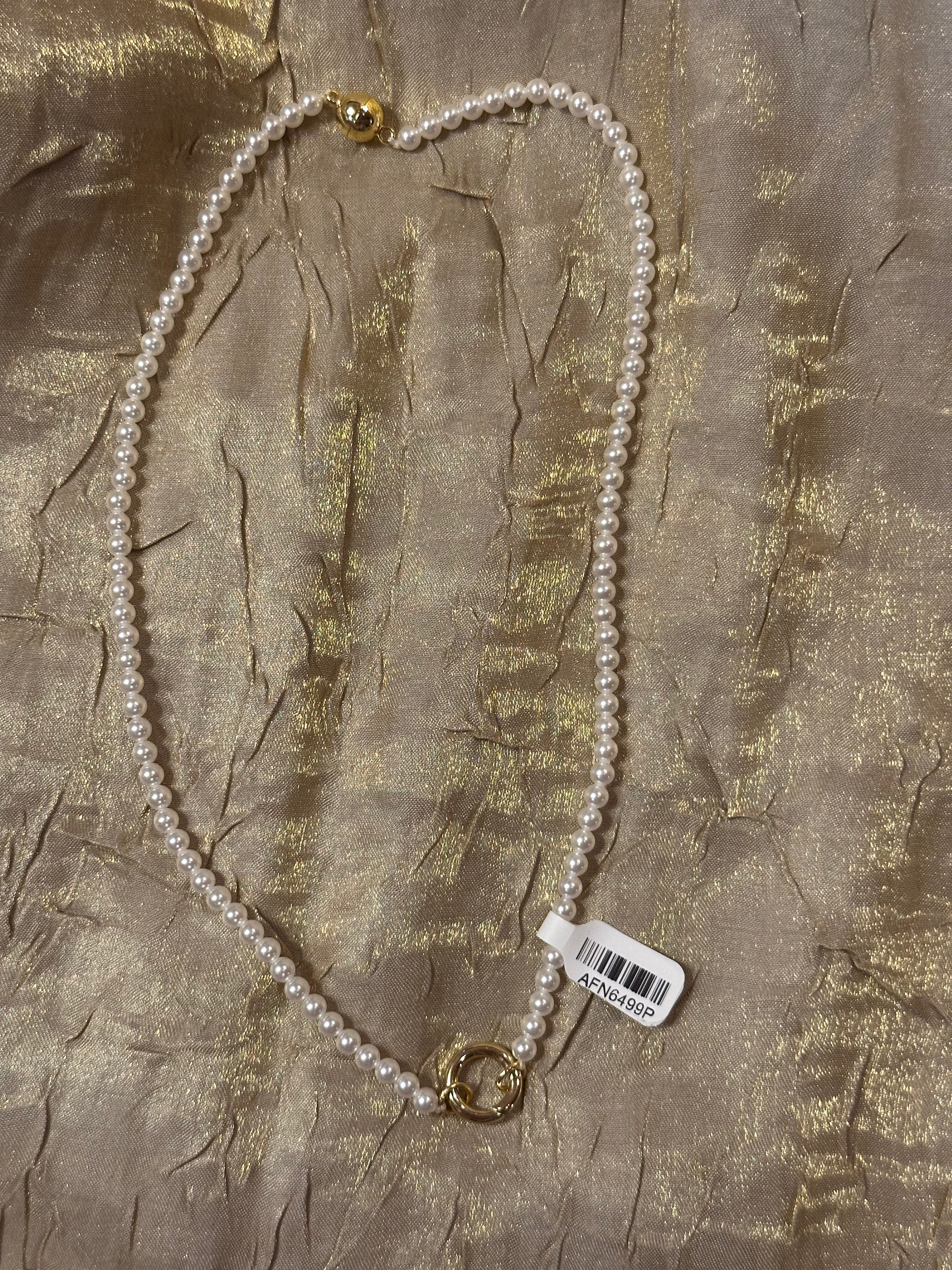 A white pearl necklace with gold-toned metal clasp details, laid flat on a textured gold fabric background with a white barcode tag attached.