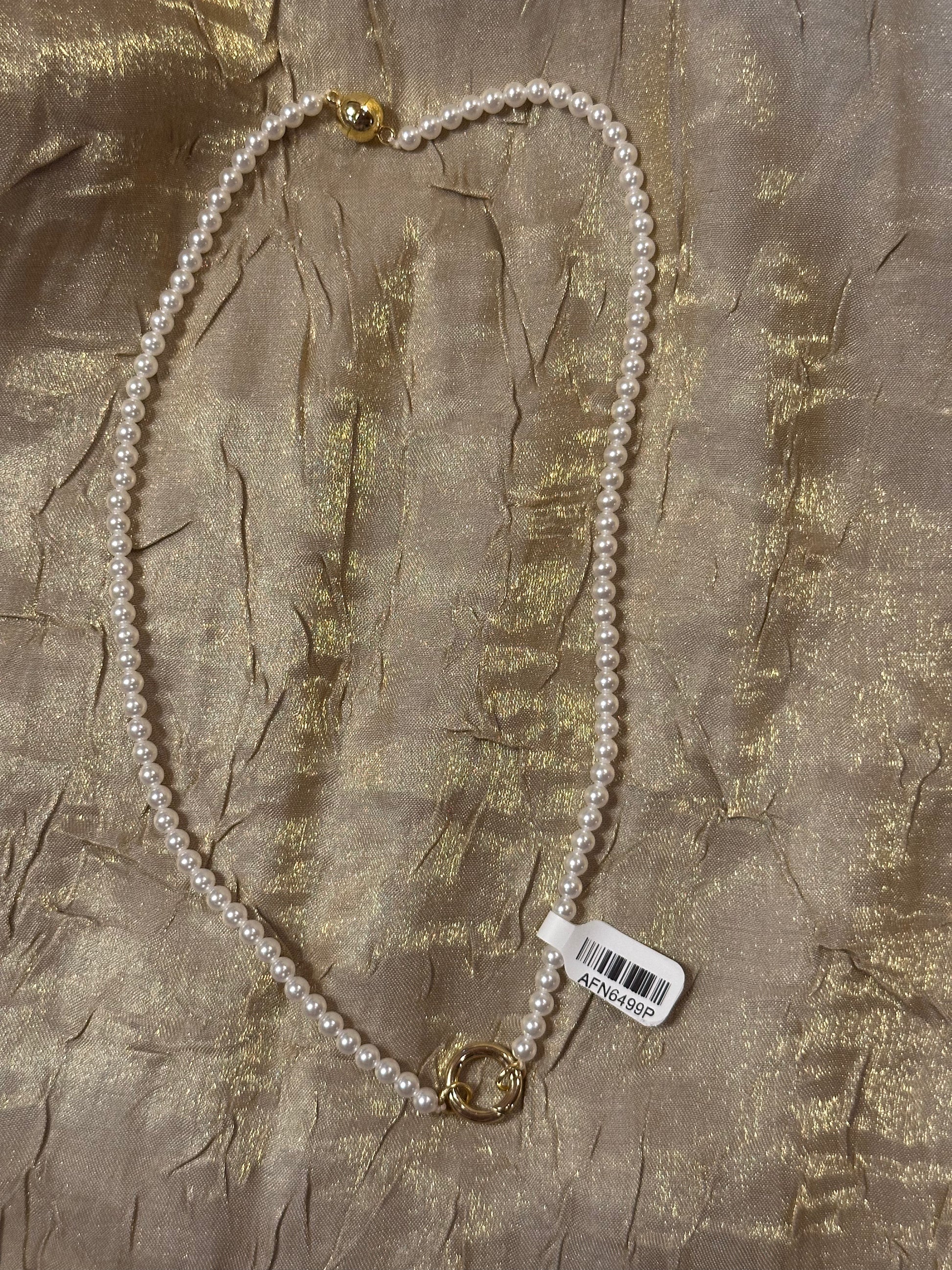 A white pearl necklace with gold-toned metal clasp details, laid flat on a textured gold fabric background with a white barcode tag attached.
