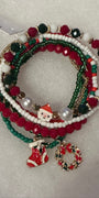 A set of five Christmas-themed beaded stretch bracelets on a white textured background. The bracelets feature red, green, and white beads, pearl-like white beads, gold-tone spacer rings, and charms including a Santa Claus figure, a red stocking, and a red and green wreath.