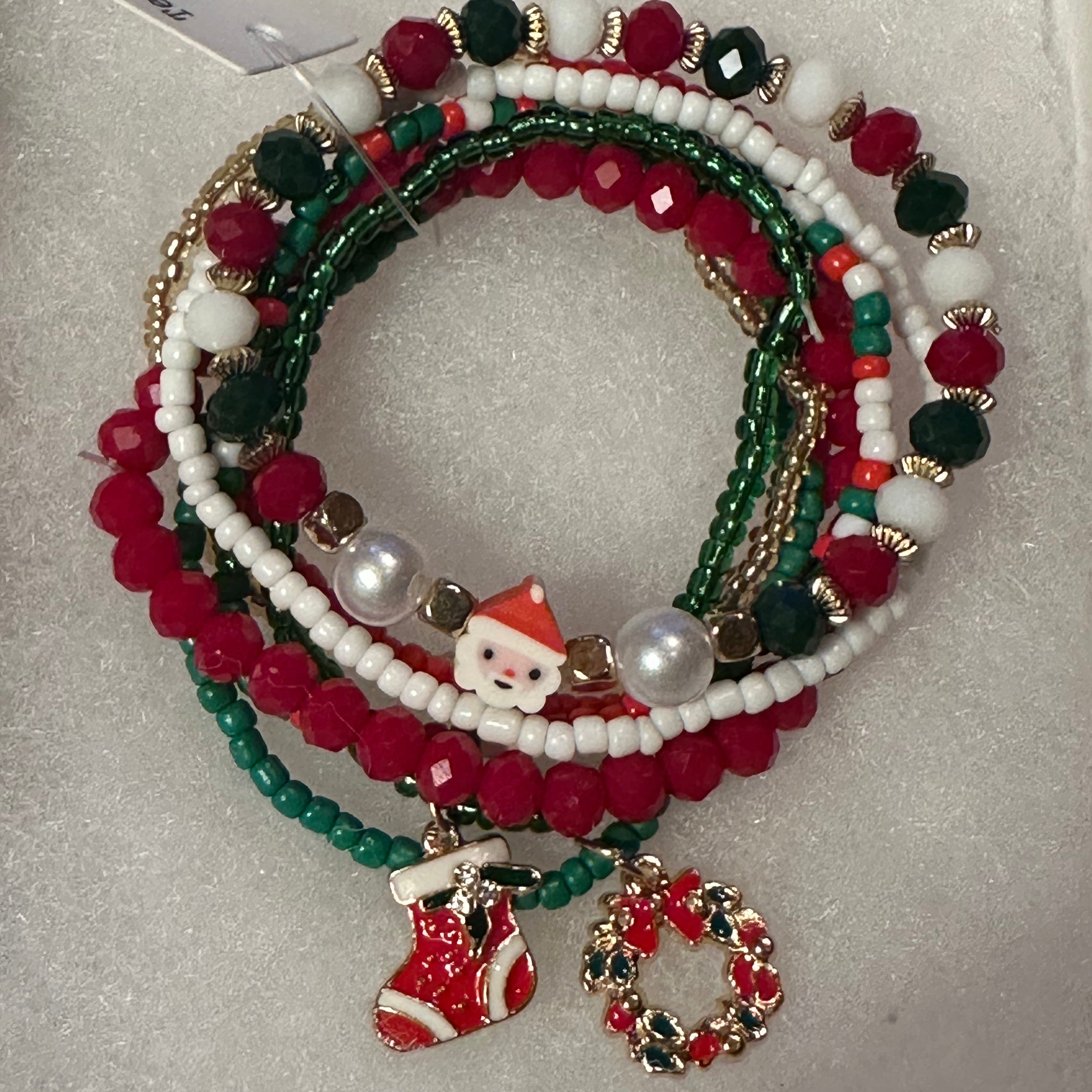 A set of five Christmas-themed beaded stretch bracelets on a white textured background. The bracelets feature red, green, and white beads, pearl-like white beads, gold-tone spacer rings, and charms including a Santa Claus figure, a red stocking, and a red and green wreath.