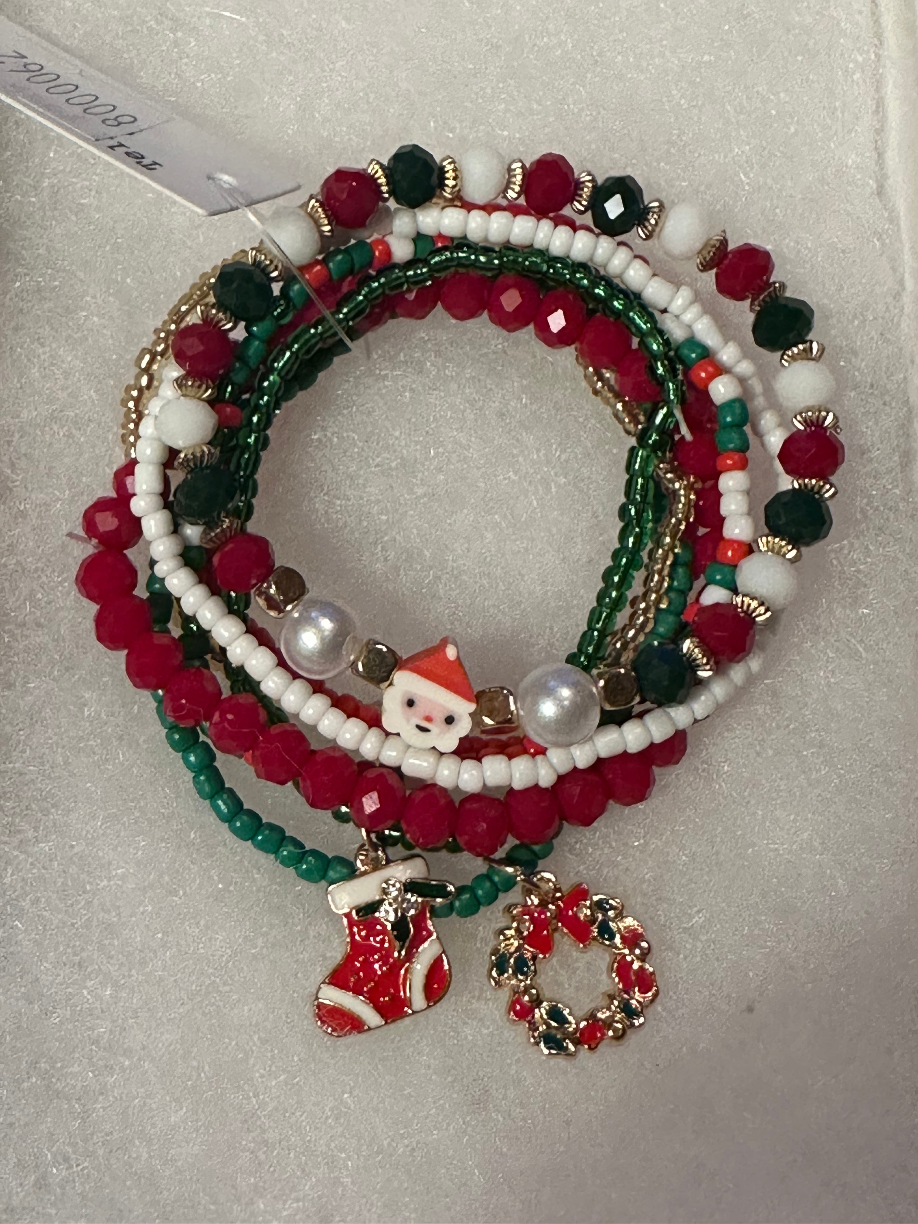 A set of five Christmas-themed beaded stretch bracelets on a white textured background. The bracelets feature red, green, and white beads, pearl-like white beads, gold-tone spacer rings, and charms including a Santa Claus figure, a red stocking, and a red and green wreath.