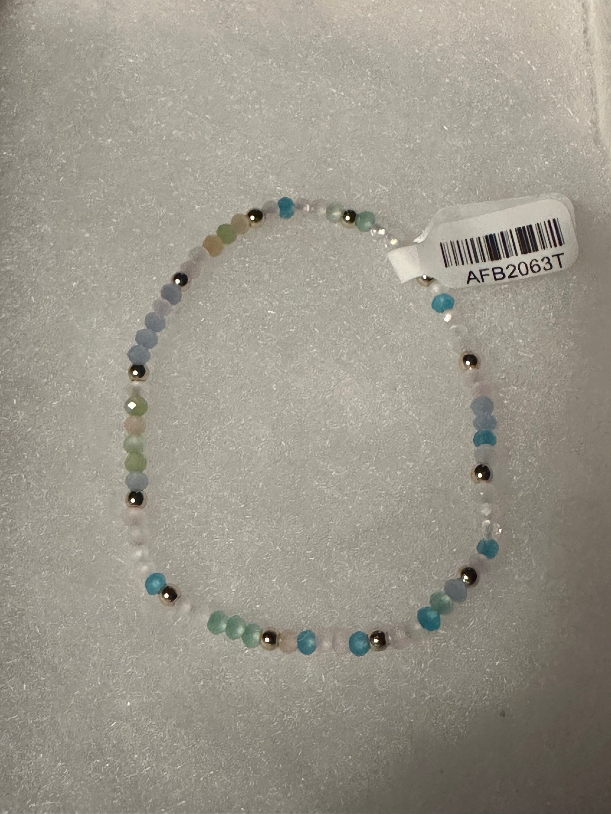 A beaded stretch bracelet made of small translucent blue, green, and white beads with a few metallic silver beads, shown on a white textured background with a white product tag attached.