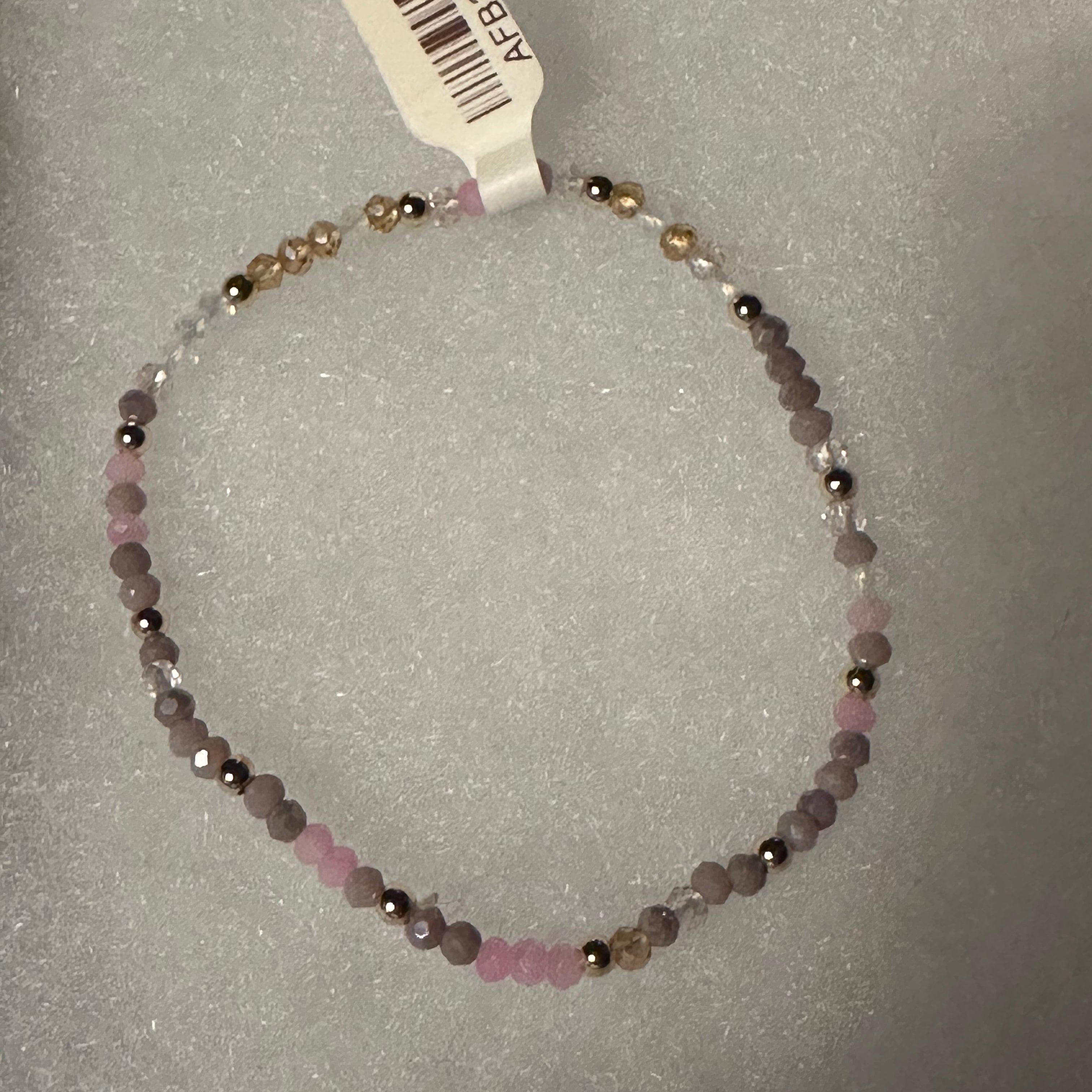 A beaded stretch bracelet with faceted pink, lavender, and metallic gold beads, shown with a white product tag on a textured gray background.