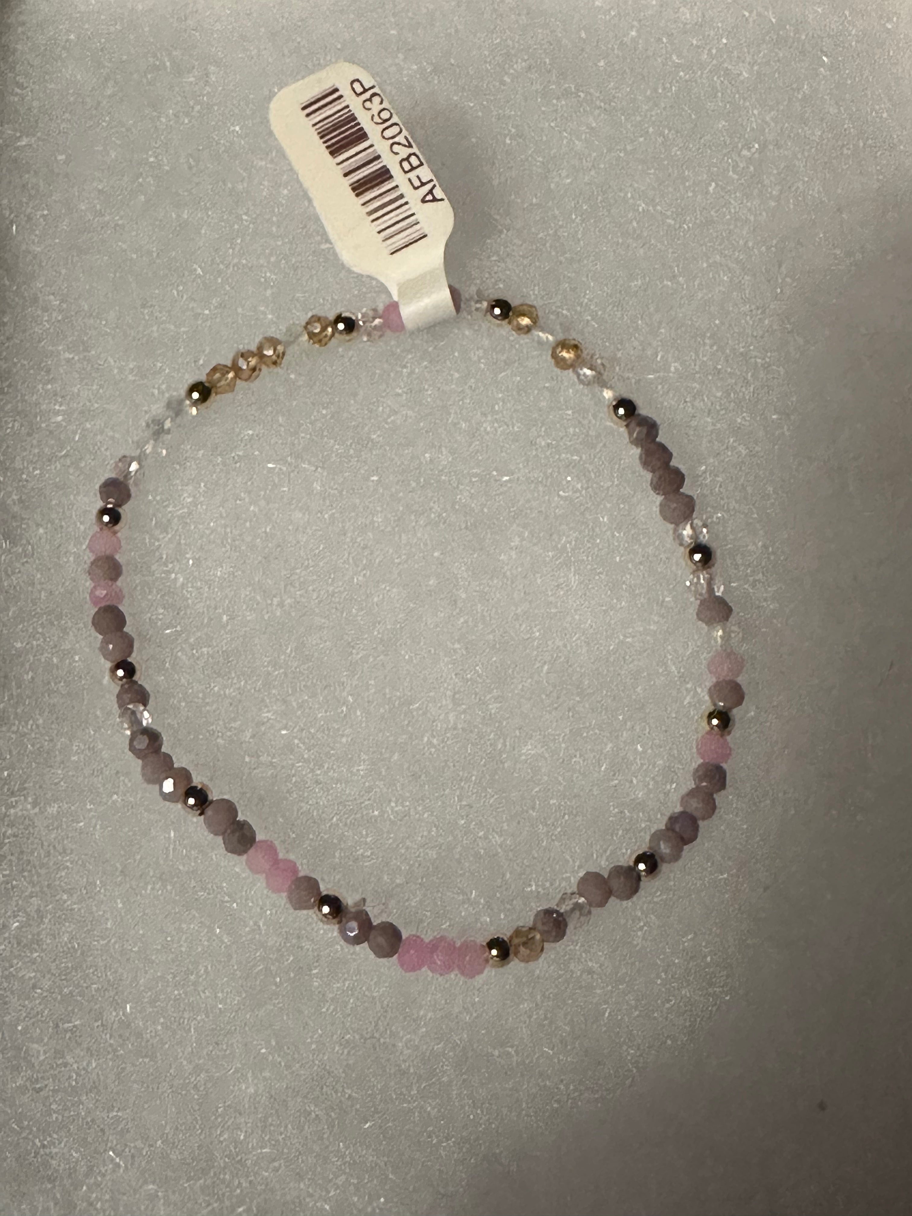 A beaded stretch bracelet with faceted pink, lavender, and metallic gold beads, shown with a white product tag on a textured gray background.