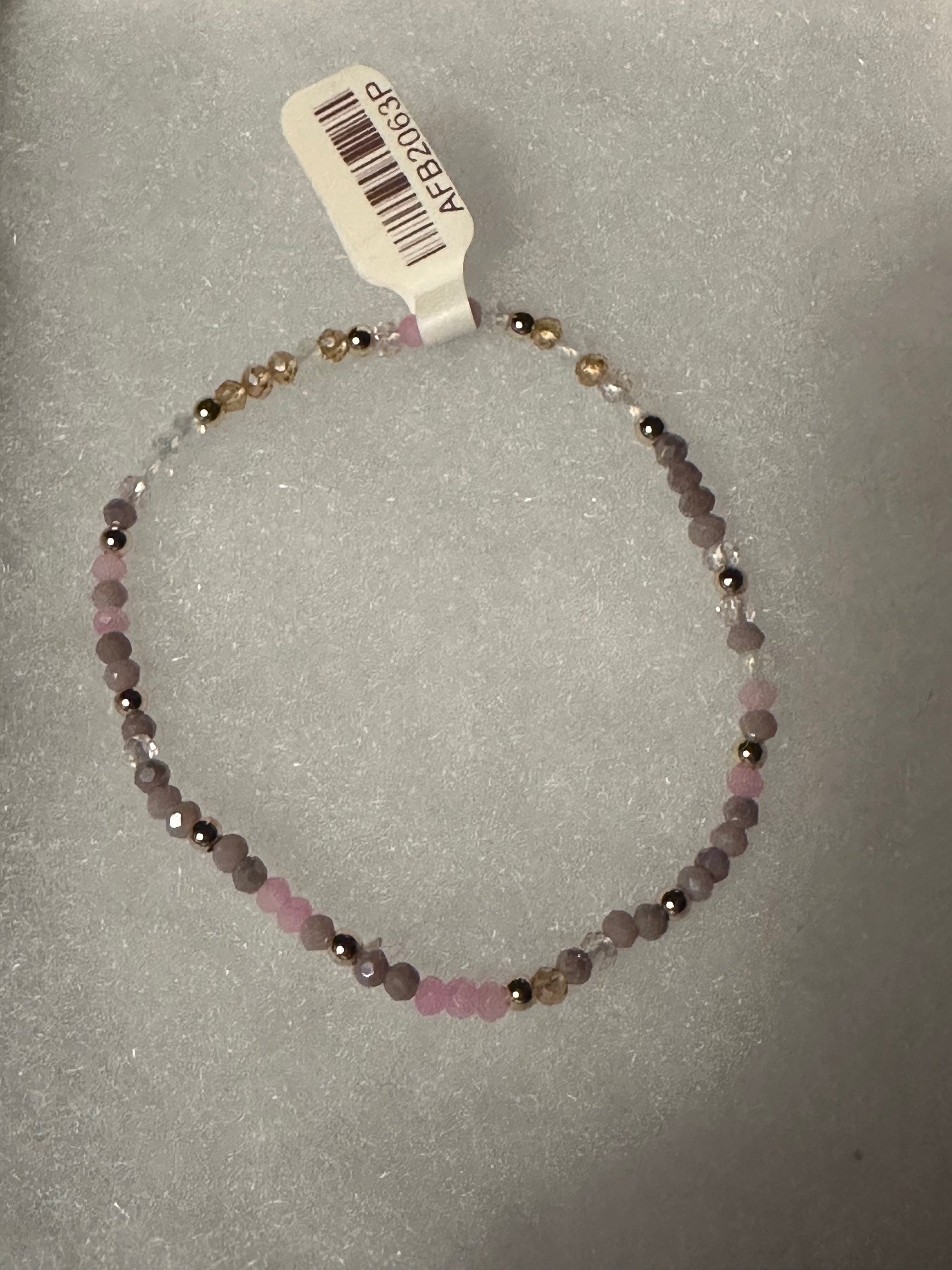 A beaded stretch bracelet with faceted pink, lavender, and metallic gold beads, shown with a white product tag on a textured gray background.