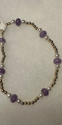 A beaded stretch bracelet featuring purple amethyst beads, white pearl-like beads, and metallic gold-toned spacer beads, shown on a textured light gray background with a white product tag attached.