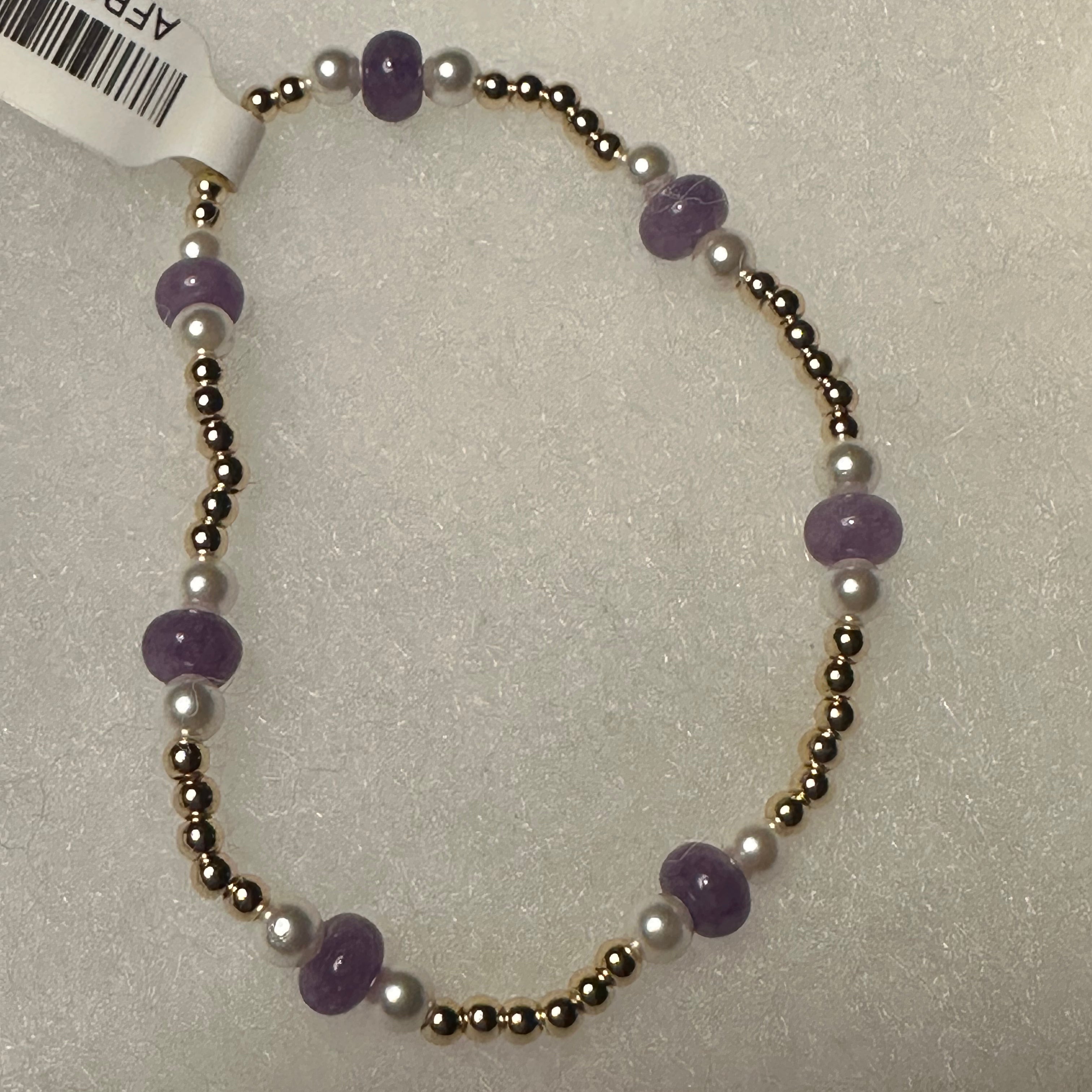 A beaded stretch bracelet featuring purple amethyst beads, white pearl-like beads, and metallic gold-toned spacer beads, shown on a textured light gray background with a white product tag attached.