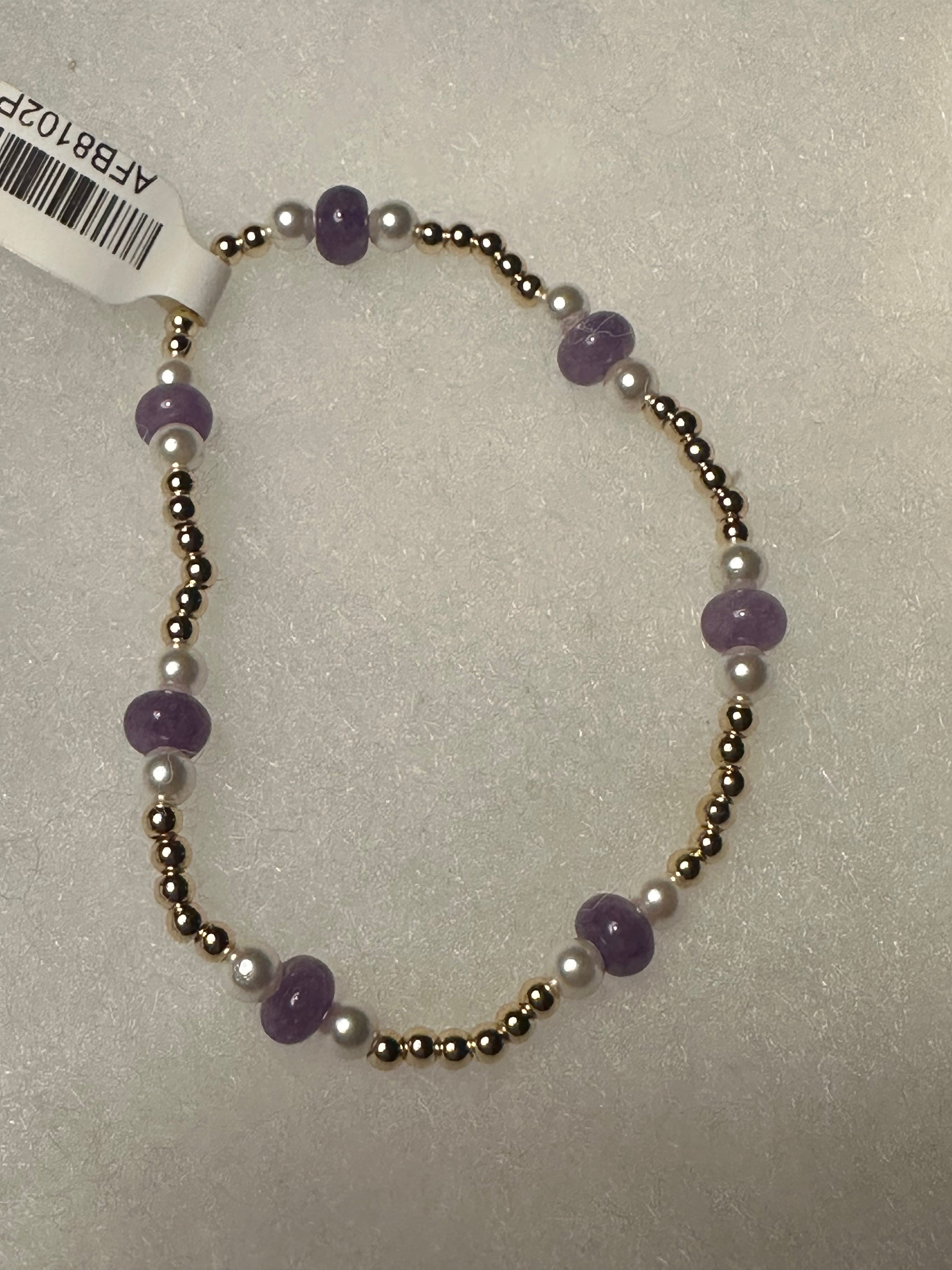 A beaded stretch bracelet featuring purple amethyst beads, white pearl-like beads, and metallic gold-toned spacer beads, shown on a textured light gray background with a white product tag attached.