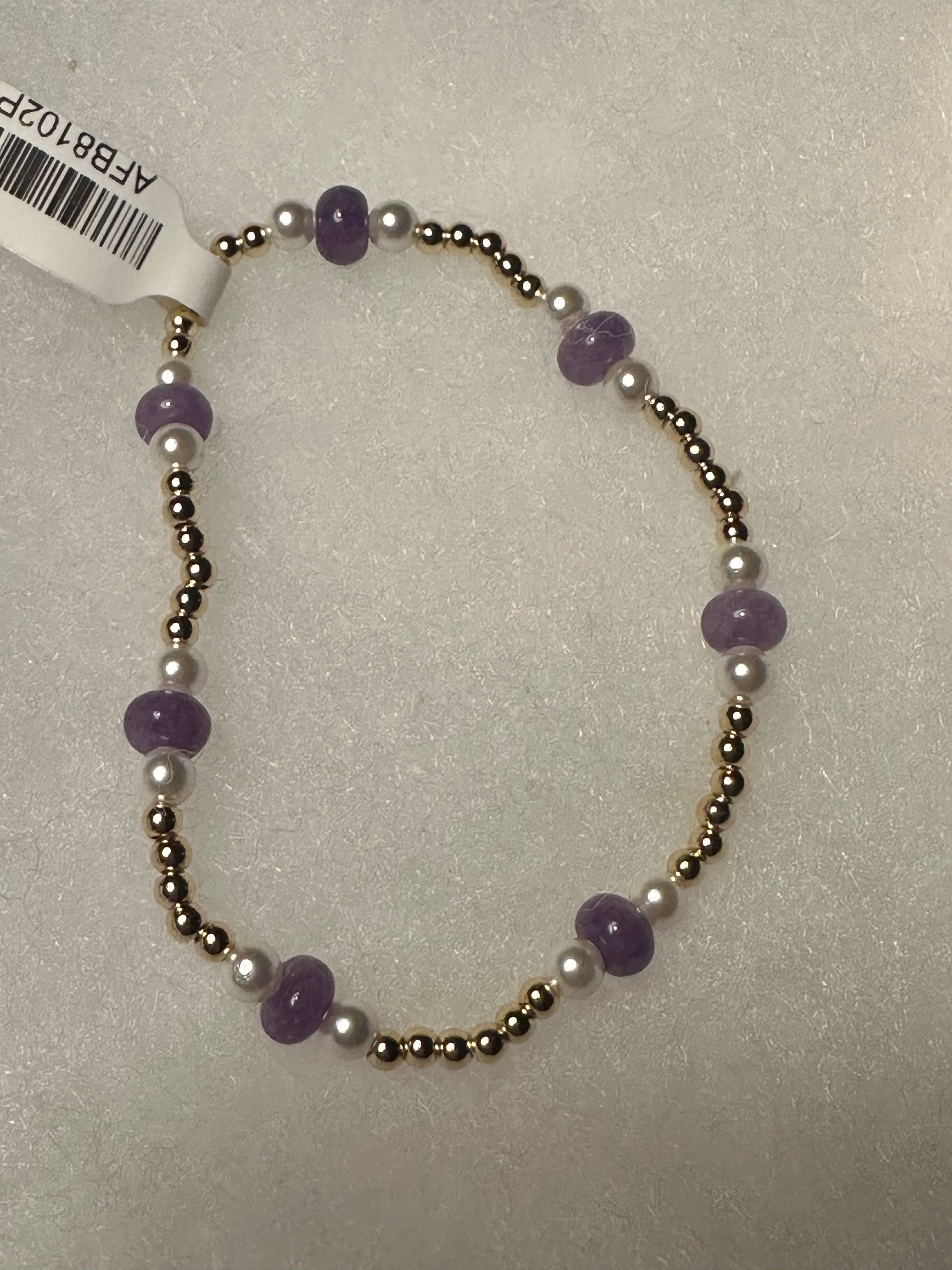A beaded stretch bracelet featuring purple amethyst beads, white pearl-like beads, and metallic gold-toned spacer beads, shown on a textured light gray background with a white product tag attached.