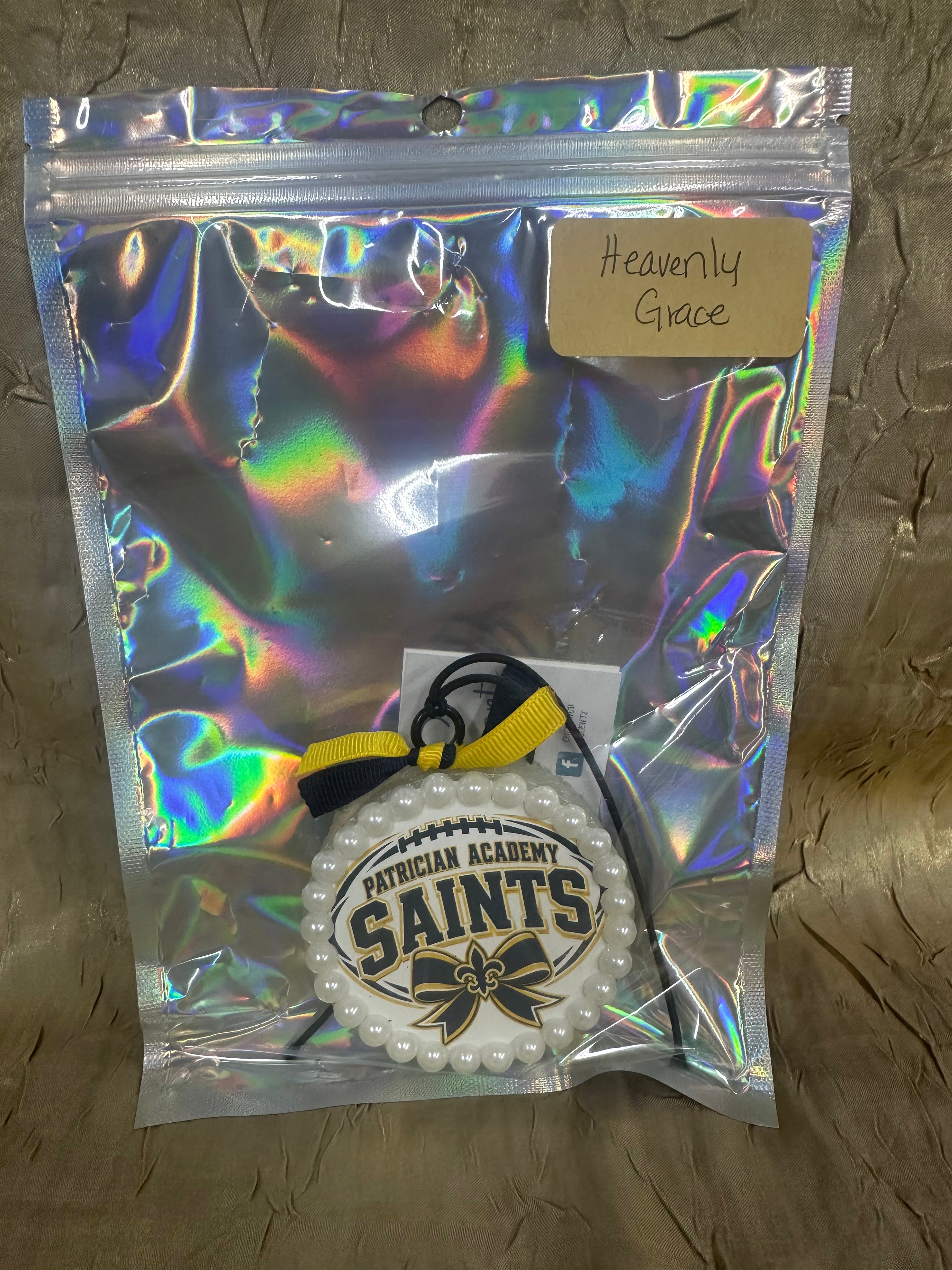 A packaged white beaded round ornament with a black and yellow ribbon and a central label reading 'Patrician Academy Saints' and 'Heavenly Grace,' sealed in an iridescent holographic bag on a textured tan surface.