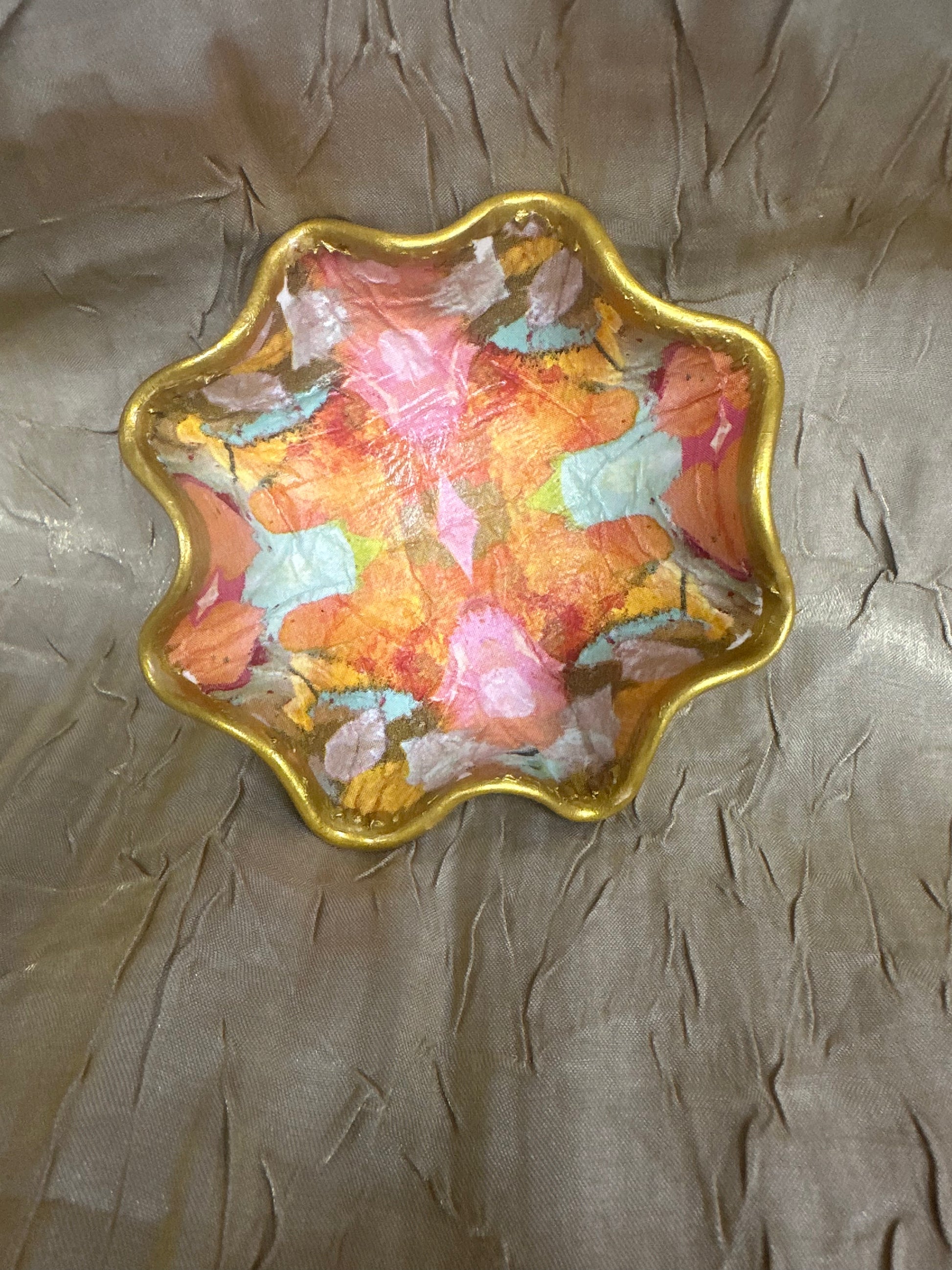 A scalloped gold metal dish with a colorful, abstract hand-painted pattern in shades of orange, pink, and blue, resting on a textured beige fabric background.