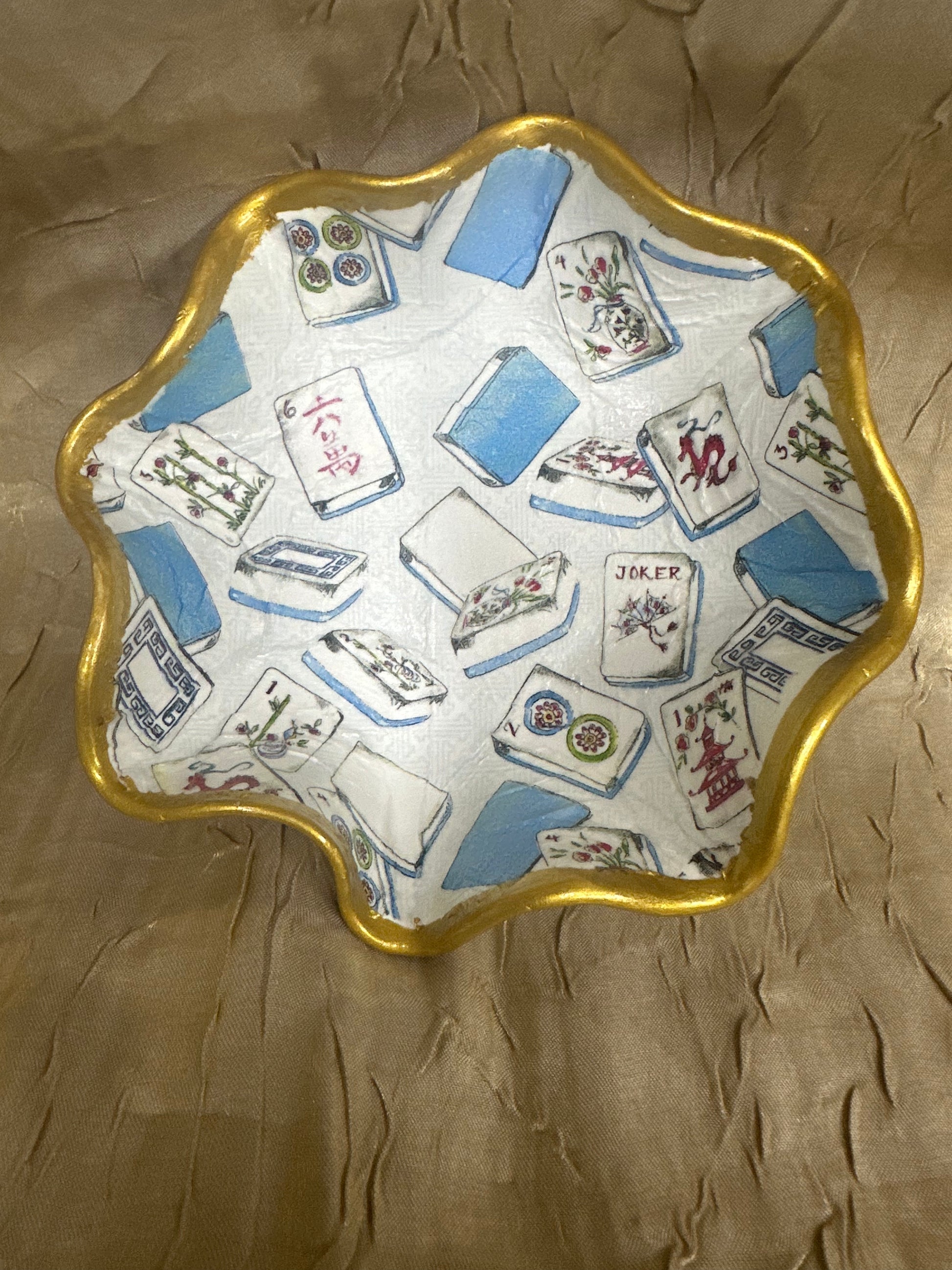 A scalloped ceramic dish featuring a white base printed with a blue and multicolor mahjong tile pattern, finished with a metallic gold rim, set against a textured brown background.