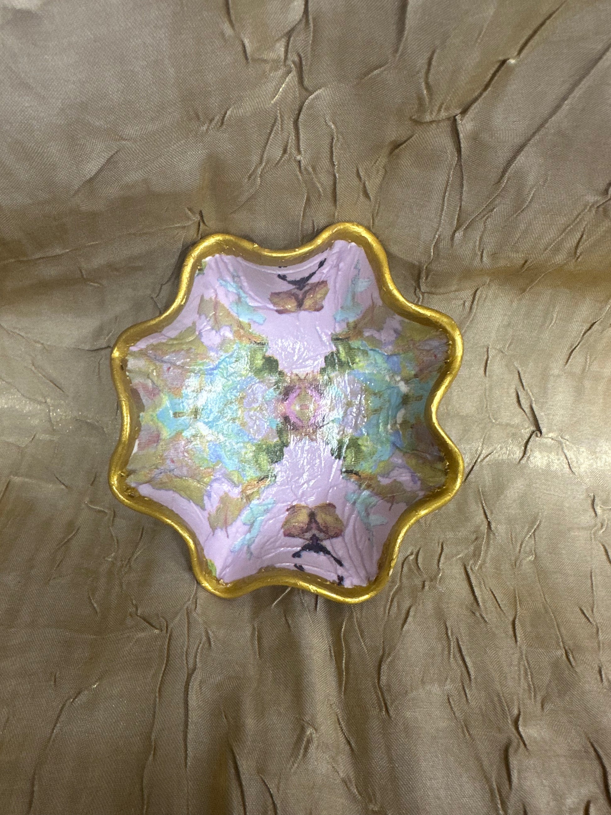 A scalloped ceramic dish with a gold rim, featuring a pastel pink and green abstract painted pattern with butterfly motifs, set against a textured gold background.