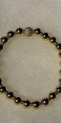 A beaded stretch bracelet featuring metallic gold and gunmetal gray round beads, with a textured silver-toned clasp, displayed on a textured white background.