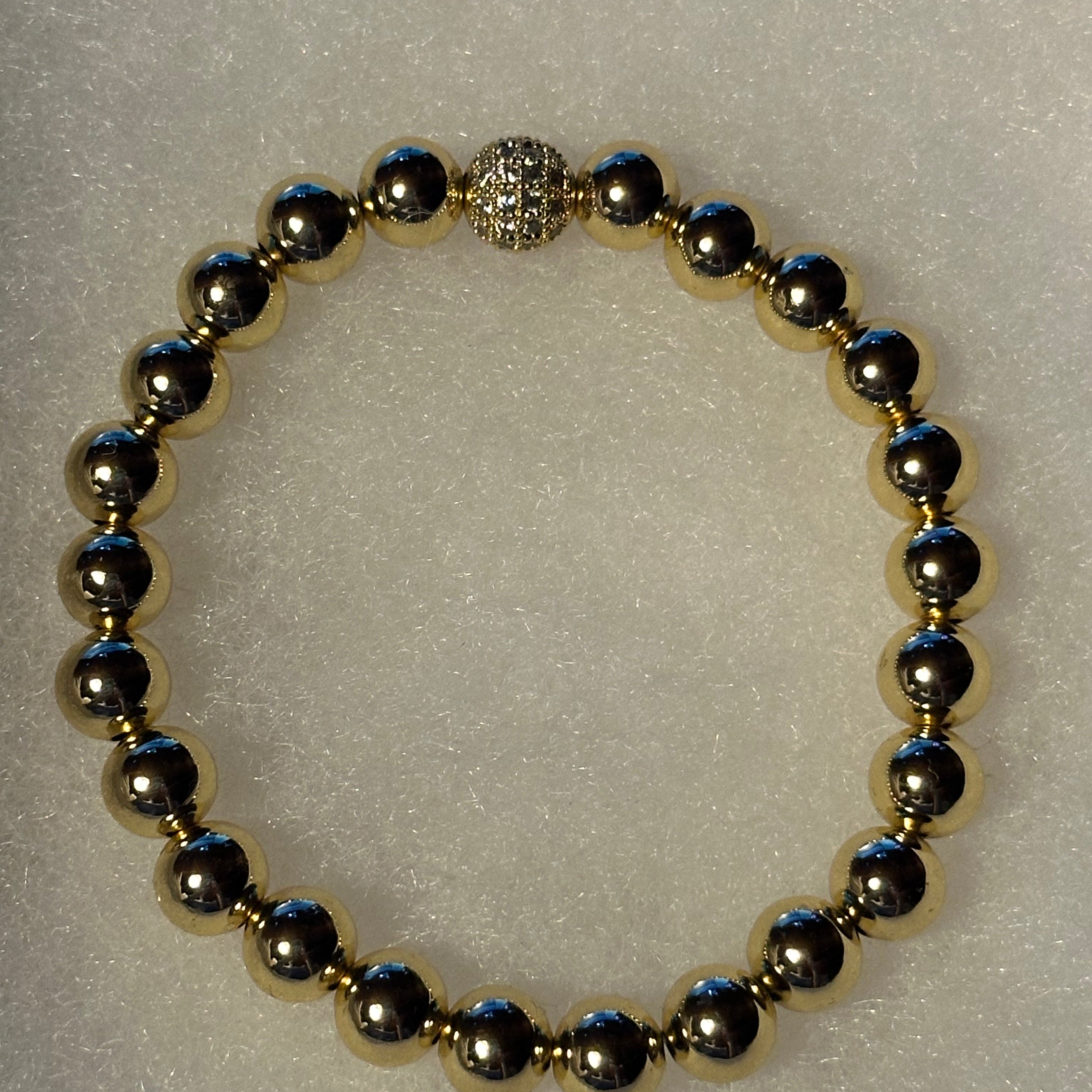 A beaded stretch bracelet featuring metallic gold and gunmetal gray round beads, with a textured silver-toned clasp, displayed on a textured white background.