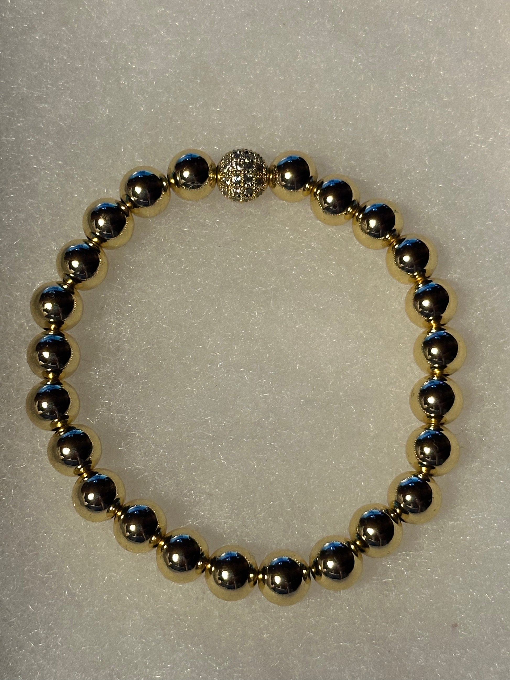 A beaded stretch bracelet featuring metallic gold and gunmetal gray round beads, with a textured silver-toned clasp, displayed on a textured white background.