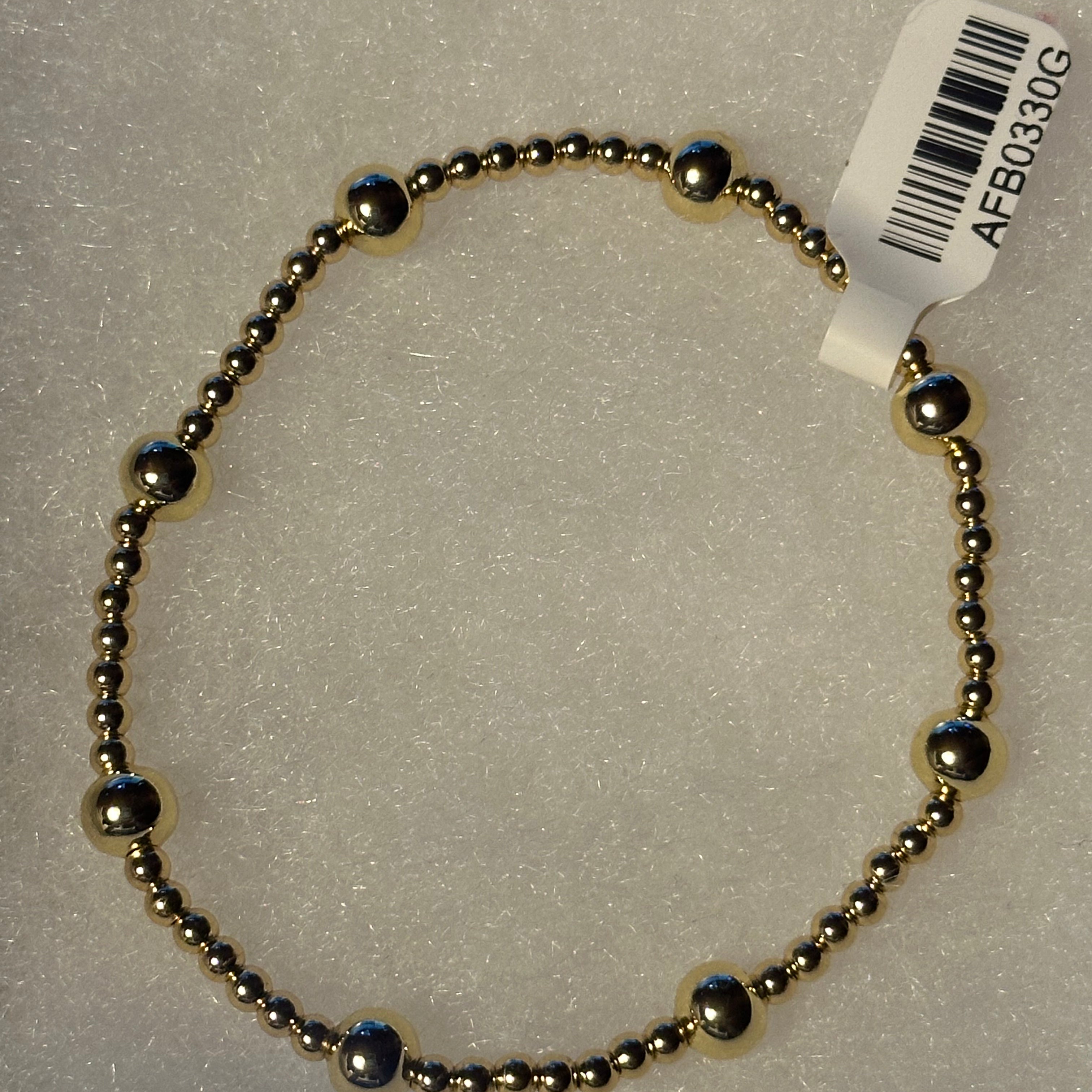 A beaded bracelet made of small metallic gold-toned beads with alternating polished silver-toned dome accents, shown on a textured white background with a white product tag attached.