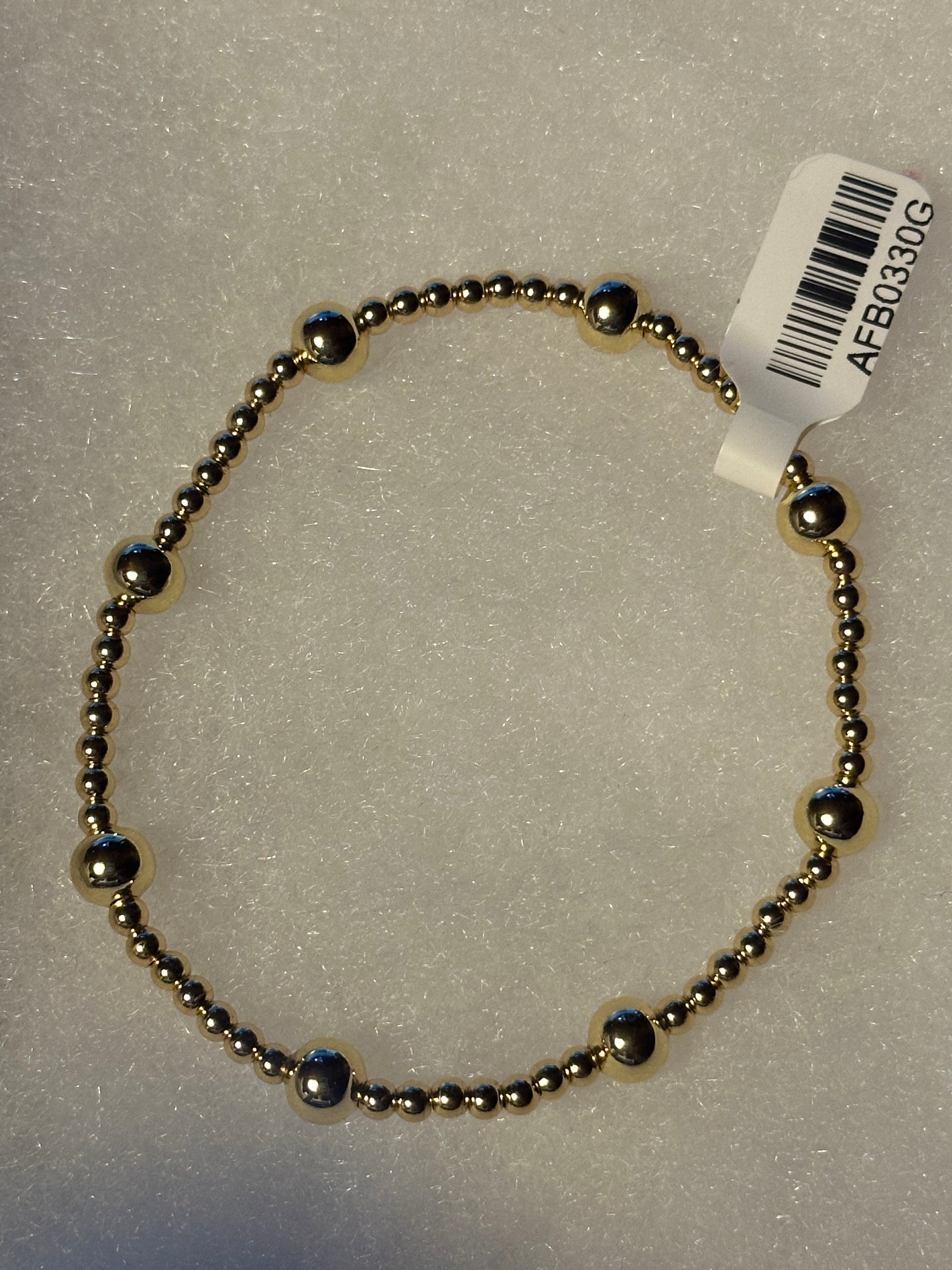 A beaded bracelet made of small metallic gold-toned beads with alternating polished silver-toned dome accents, shown on a textured white background with a white product tag attached.