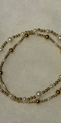 A double-strand beaded necklace featuring white faux pearls and polished gold-tone metal beads, arranged in a loose circle on a textured off-white background.