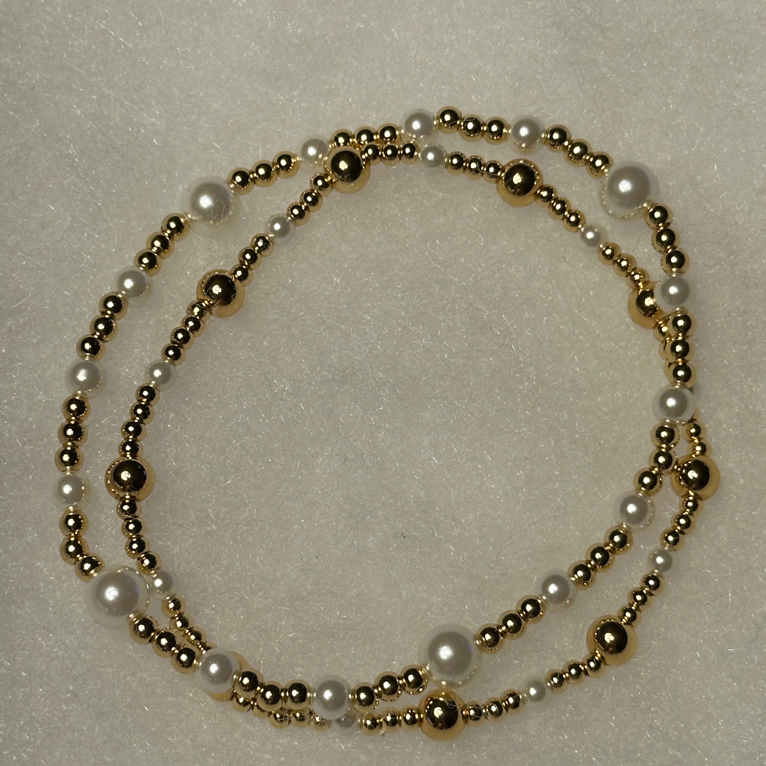 A double-strand beaded necklace featuring white faux pearls and polished gold-tone metal beads, arranged in a loose circle on a textured off-white background.