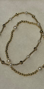 A beaded necklace featuring metallic gold-toned round beads and white faux-pearl accents, arranged in a loose loop on a textured off-white background.