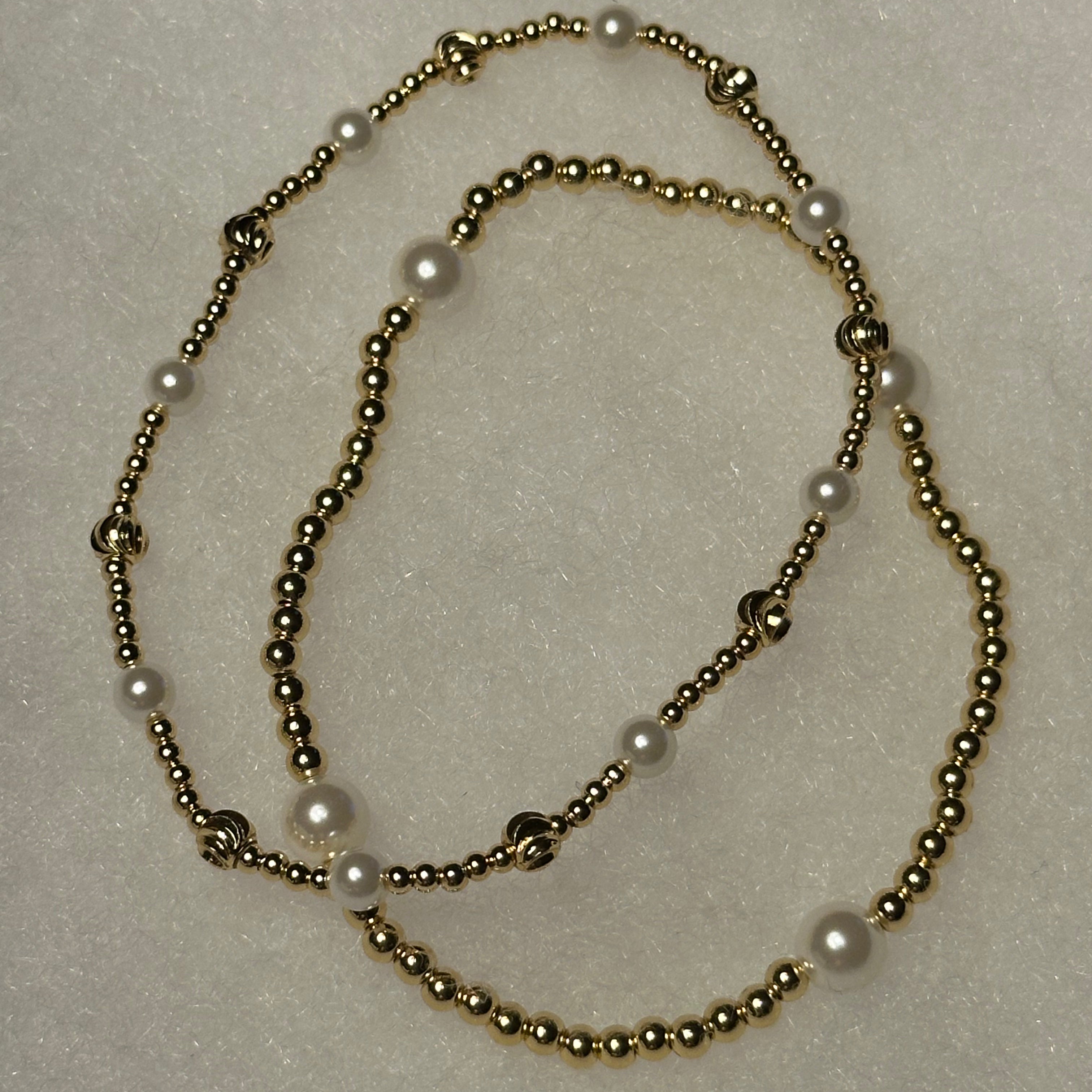 A beaded necklace featuring metallic gold-toned round beads and white faux-pearl accents, arranged in a loose loop on a textured off-white background.