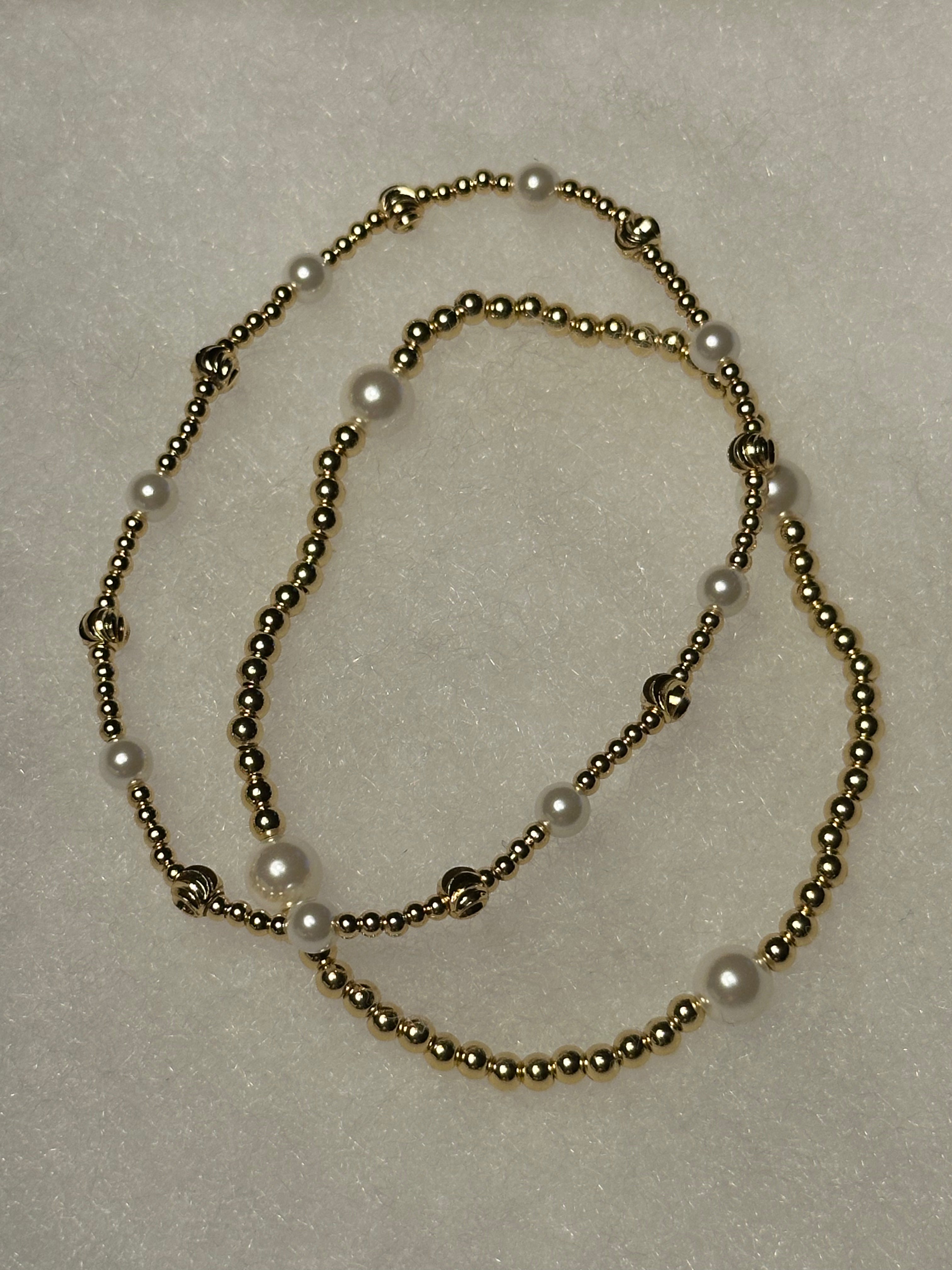 A beaded necklace featuring metallic gold-toned round beads and white faux-pearl accents, arranged in a loose loop on a textured off-white background.
