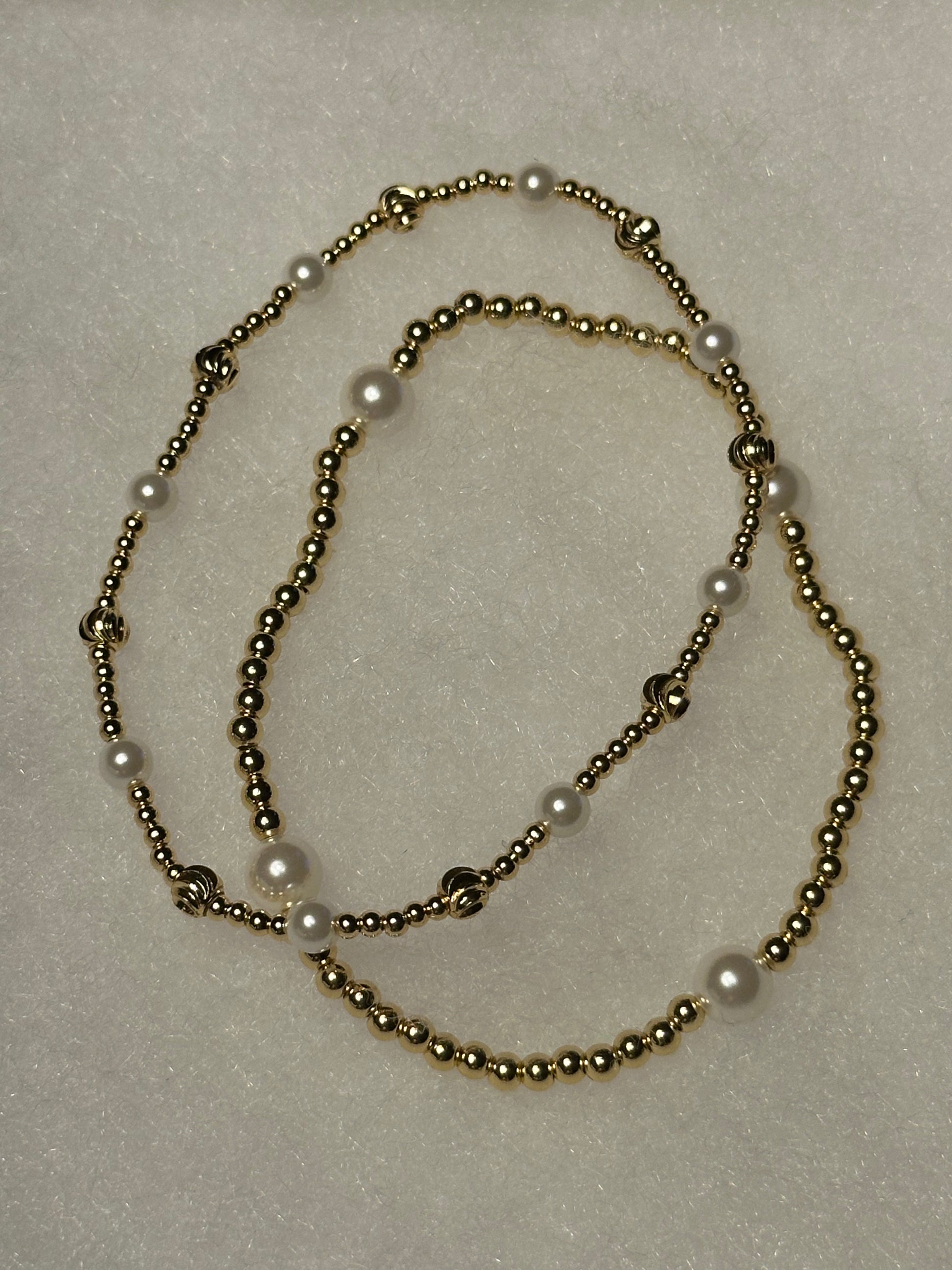 A beaded necklace featuring metallic gold-toned round beads and white faux-pearl accents, arranged in a loose loop on a textured off-white background.