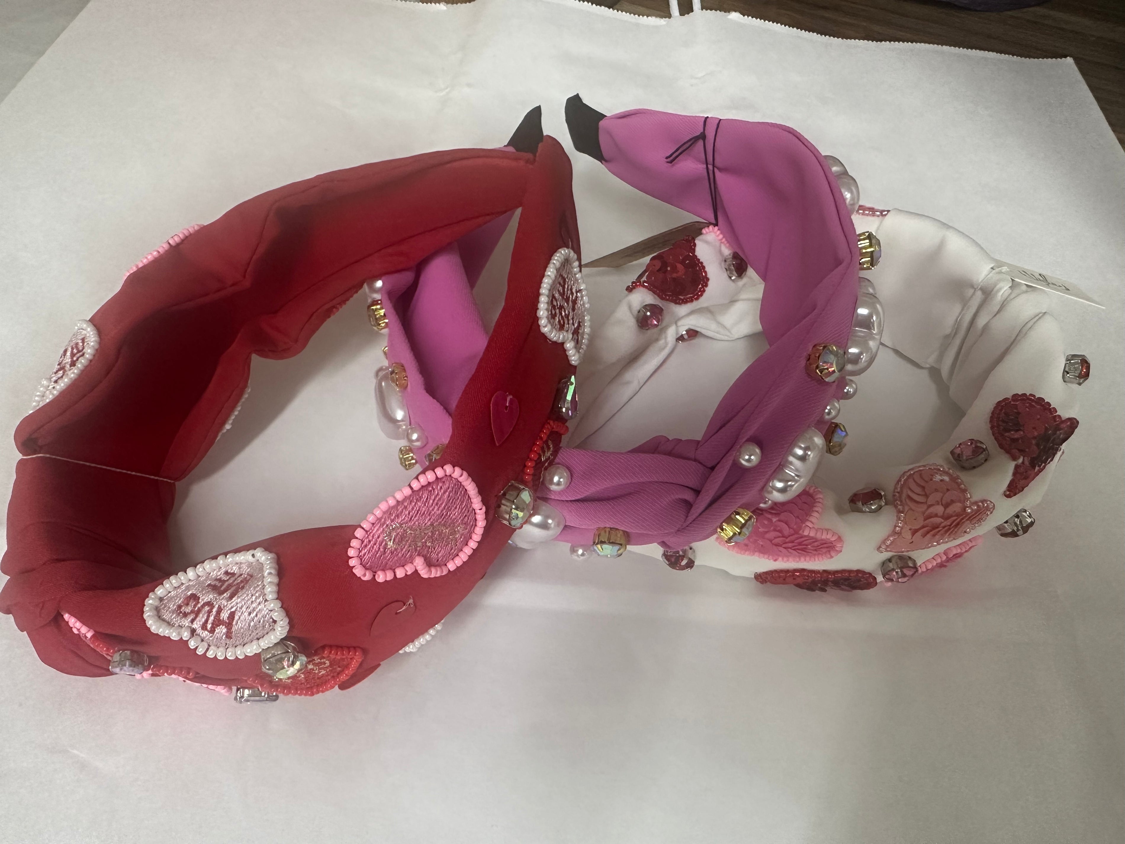 Three decorative headbands, one red . one white and one pink with heart and peaarl designs, on a white surface.