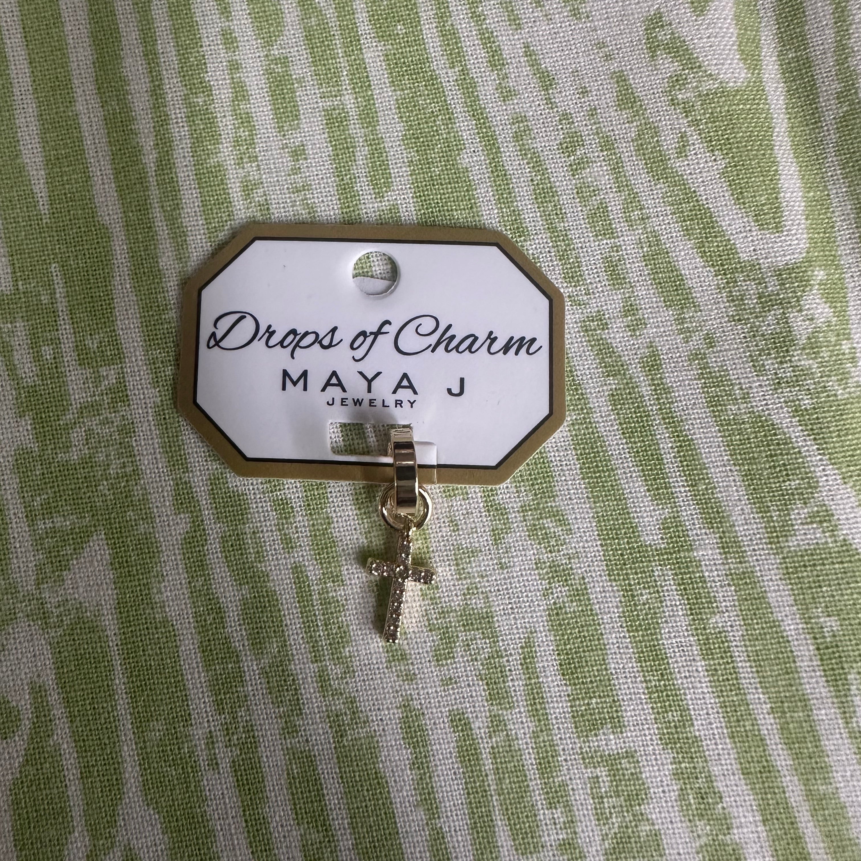 Yellow gold cross charm with 'Drops of Charm' label on a textured green background