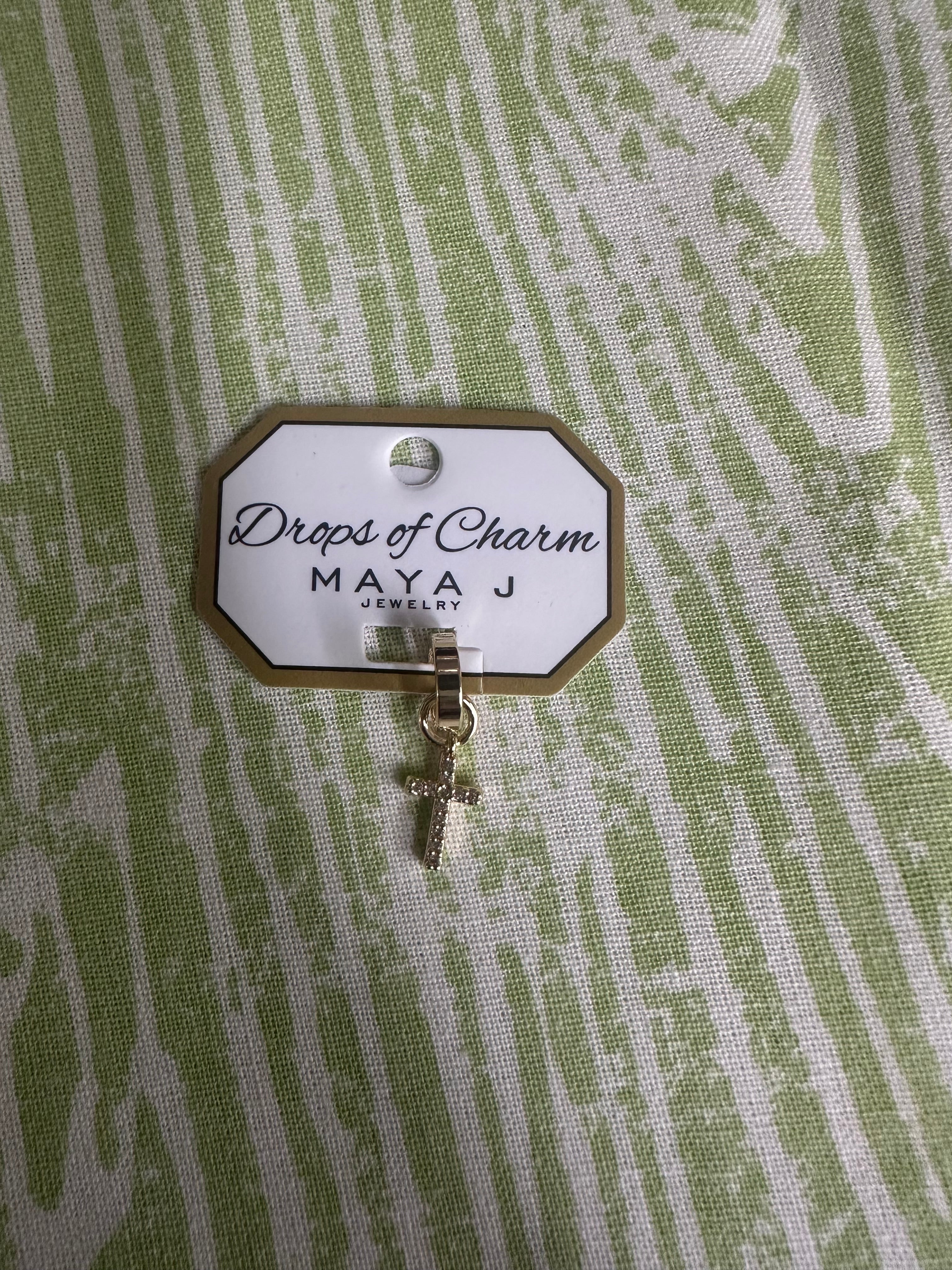 Yellow gold cross charm with 'Drops of Charm' label on a textured green background