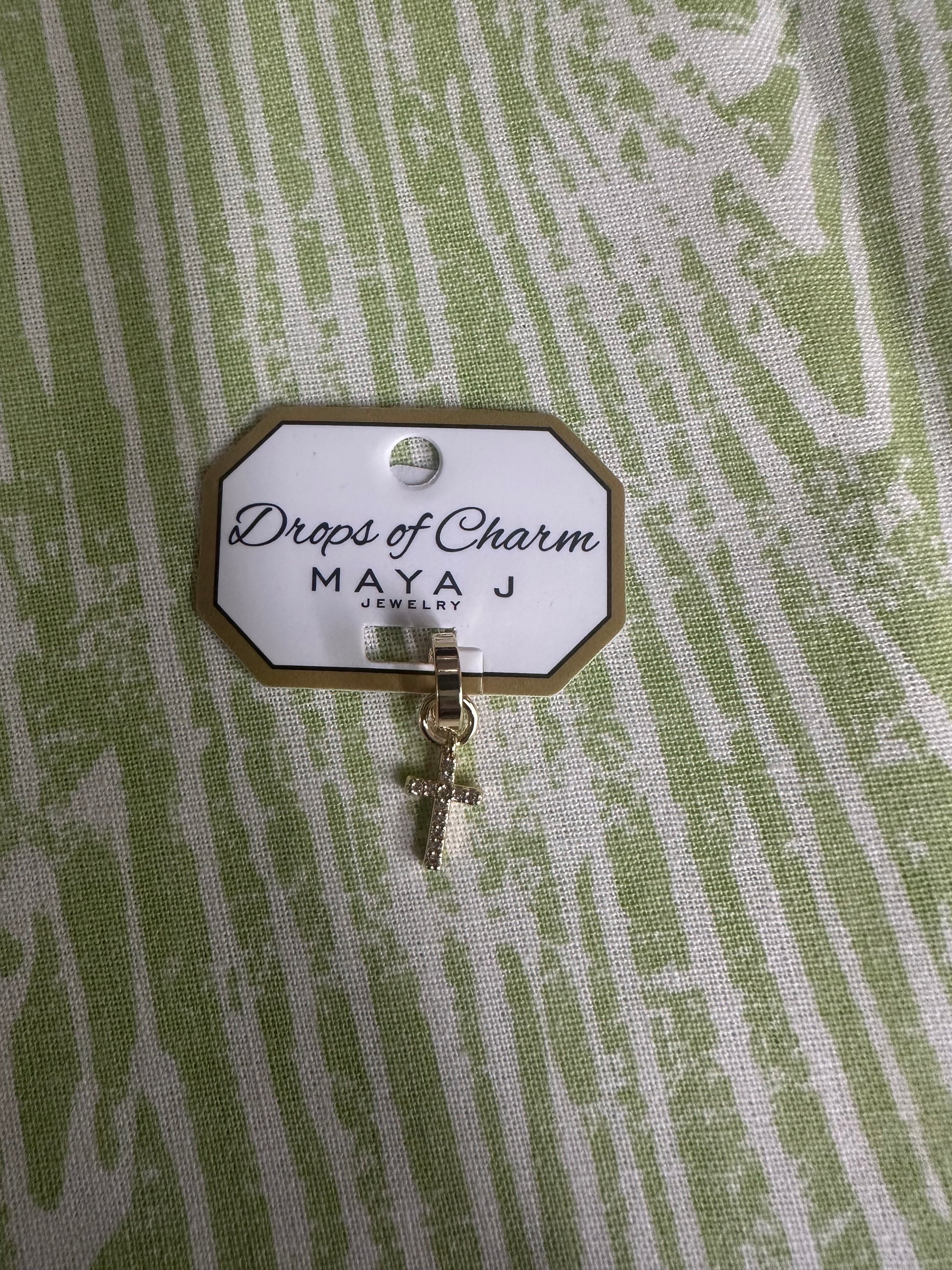 Yellow gold cross charm with 'Drops of Charm' label on a textured green background