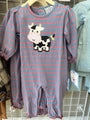 A baby long-sleeve footless romper with horizontal blue-gray and pink stripes, featuring a black-and-white cow applique on the chest, hanging on a wooden hanger on a retail display.