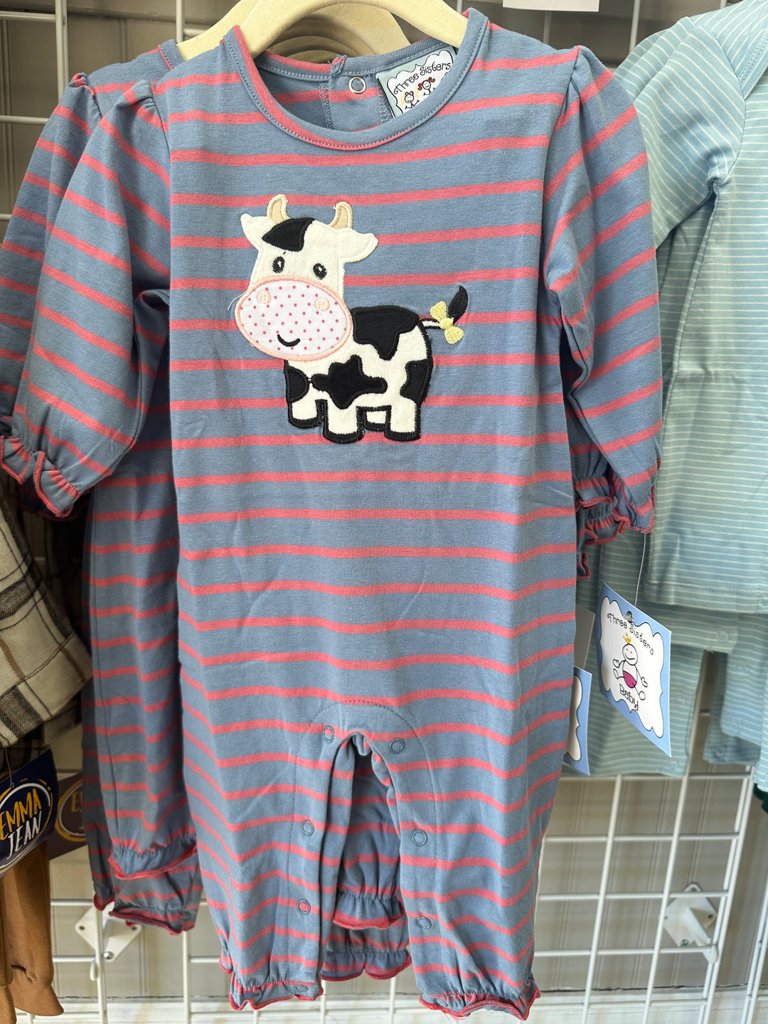 A baby long-sleeve footless romper with horizontal blue-gray and pink stripes, featuring a black-and-white cow applique on the chest, hanging on a wooden hanger on a retail display.