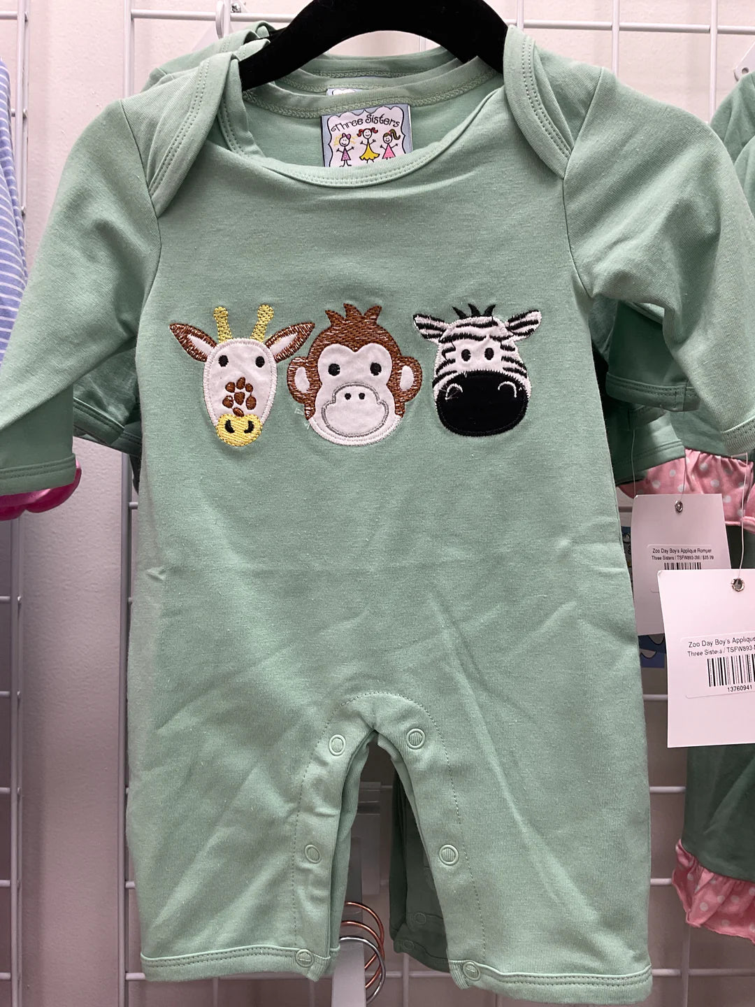 A mint green baby romper with three embroidered animal faces (giraffe, monkey, zebra) on the chest, hanging on a black hanger against a white wire display wall.
