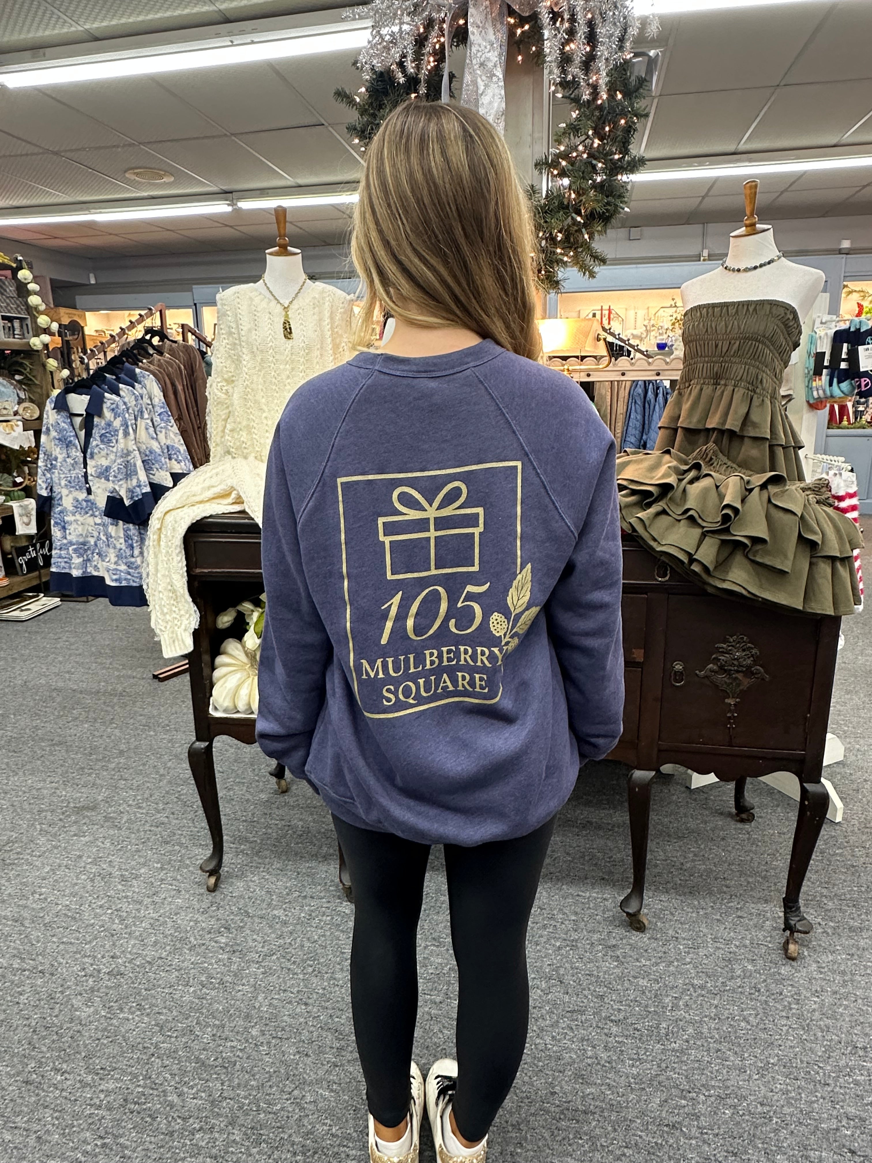 Person wearing a blue sweatshirt with a logo and text, standing in a store with racks of clothes and a mirror.