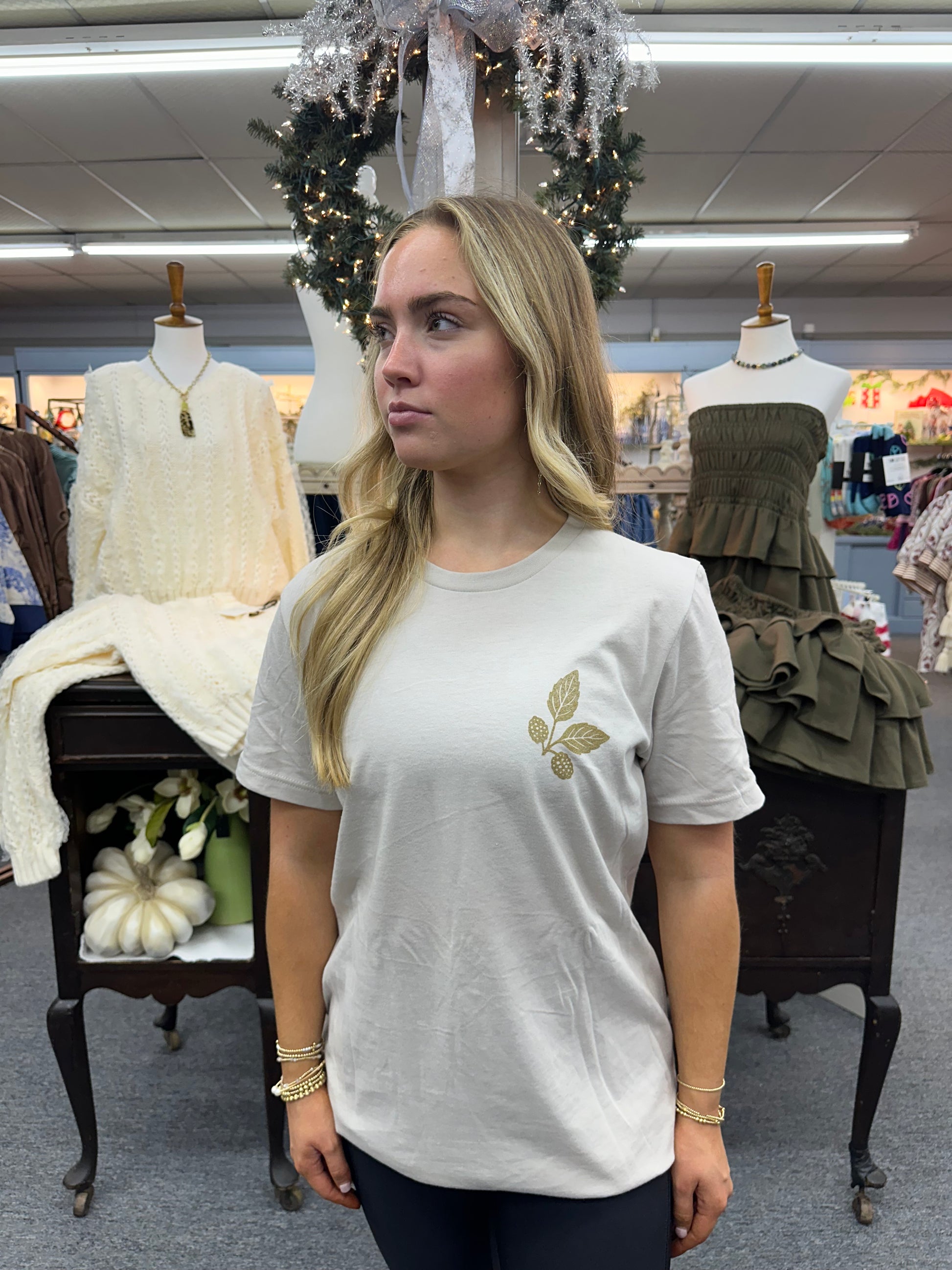 Woman wearing a sand colored t-shirt with a gold leaf design in a store setting.