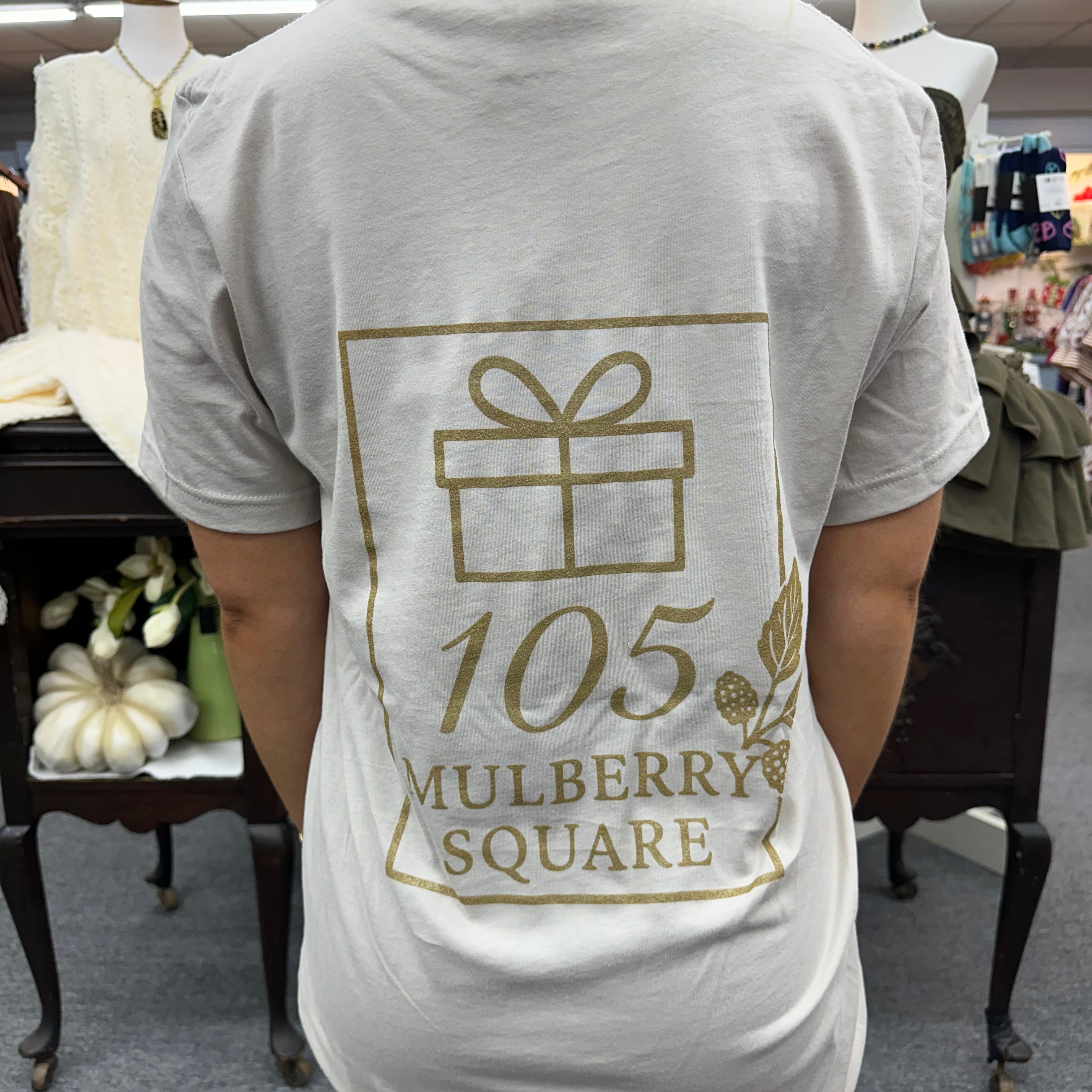 Person wearing a sand colored t-shirt with 105 Mulberry Square' printed on it, standing in a room.