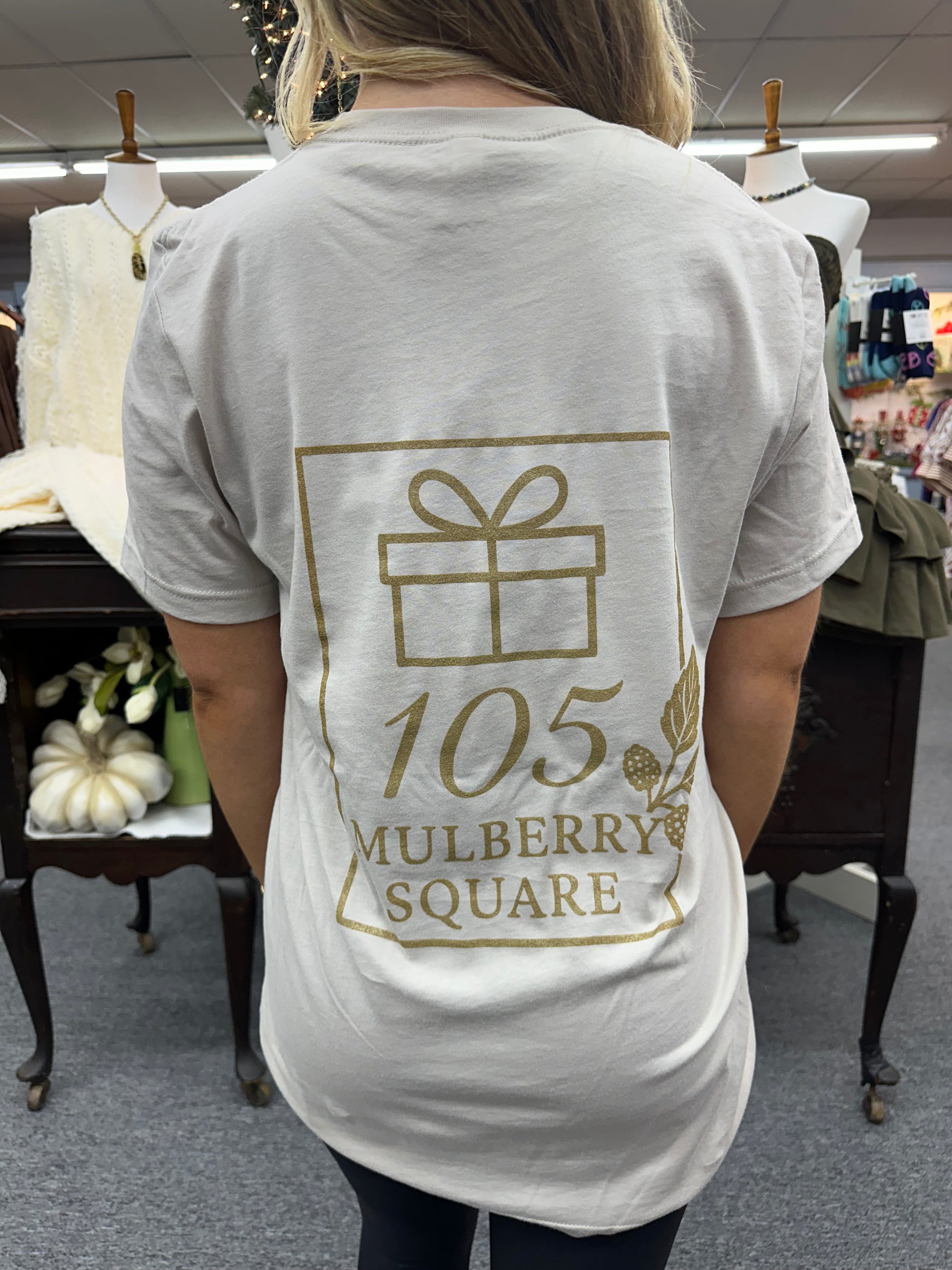 Person wearing a sand colored t-shirt with 105 Mulberry Square' printed on it, standing in a room.
