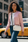 Woman in a pink bomber jacket and blue jeans standing on a city street with a yellow taxi in the background.