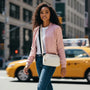 Woman in a pink bomber jacket and blue jeans standing on a city street with a yellow taxi in the background.