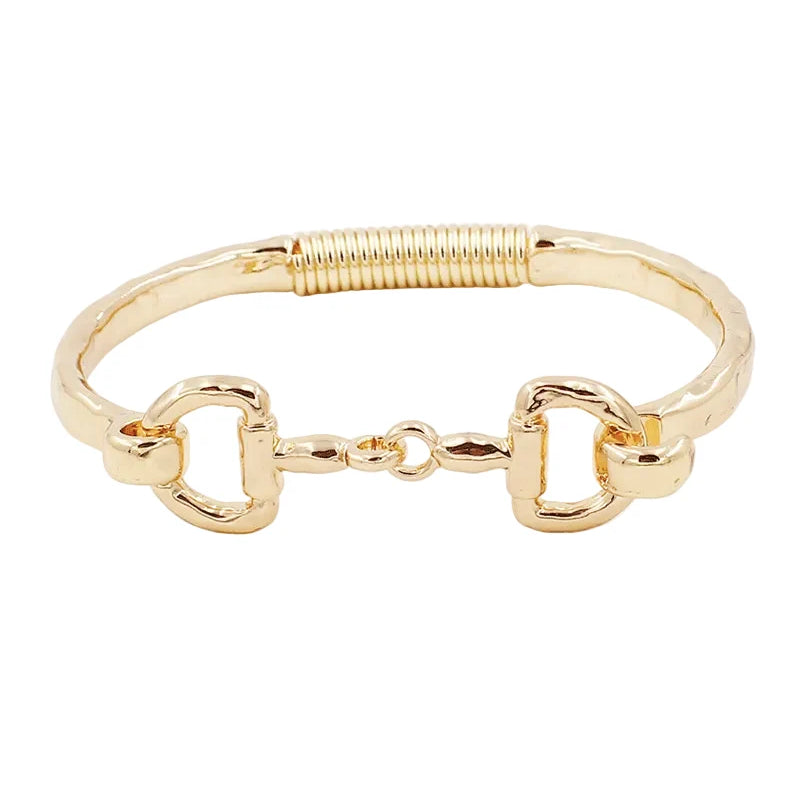 A gold-tone metal bracelet featuring a textured band, a coiled spring detail on one side, and two horsebit-style D-rings connected by a small central link, shown on a plain white background.