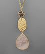 A gold-tone dangle necklace featuring a textured hammered oval pendant, a small round bezel-set cluster of dark brown stones, and a large teardrop-shaped pale pink stone pendant, shown against a solid gray background.