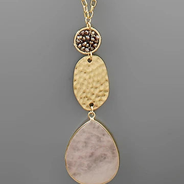 A gold-tone dangle necklace featuring a textured hammered oval pendant, a small round bezel-set cluster of dark brown stones, and a large teardrop-shaped pale pink stone pendant, shown against a solid gray background.