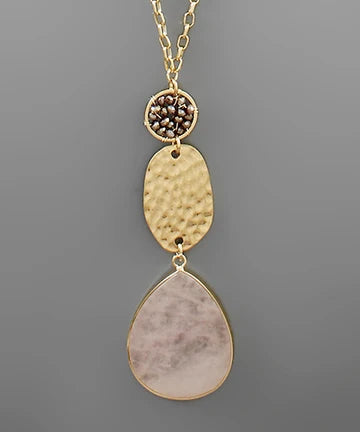 A gold-tone dangle necklace featuring a textured hammered oval pendant, a small round bezel-set cluster of dark brown stones, and a large teardrop-shaped pale pink stone pendant, shown against a solid gray background.